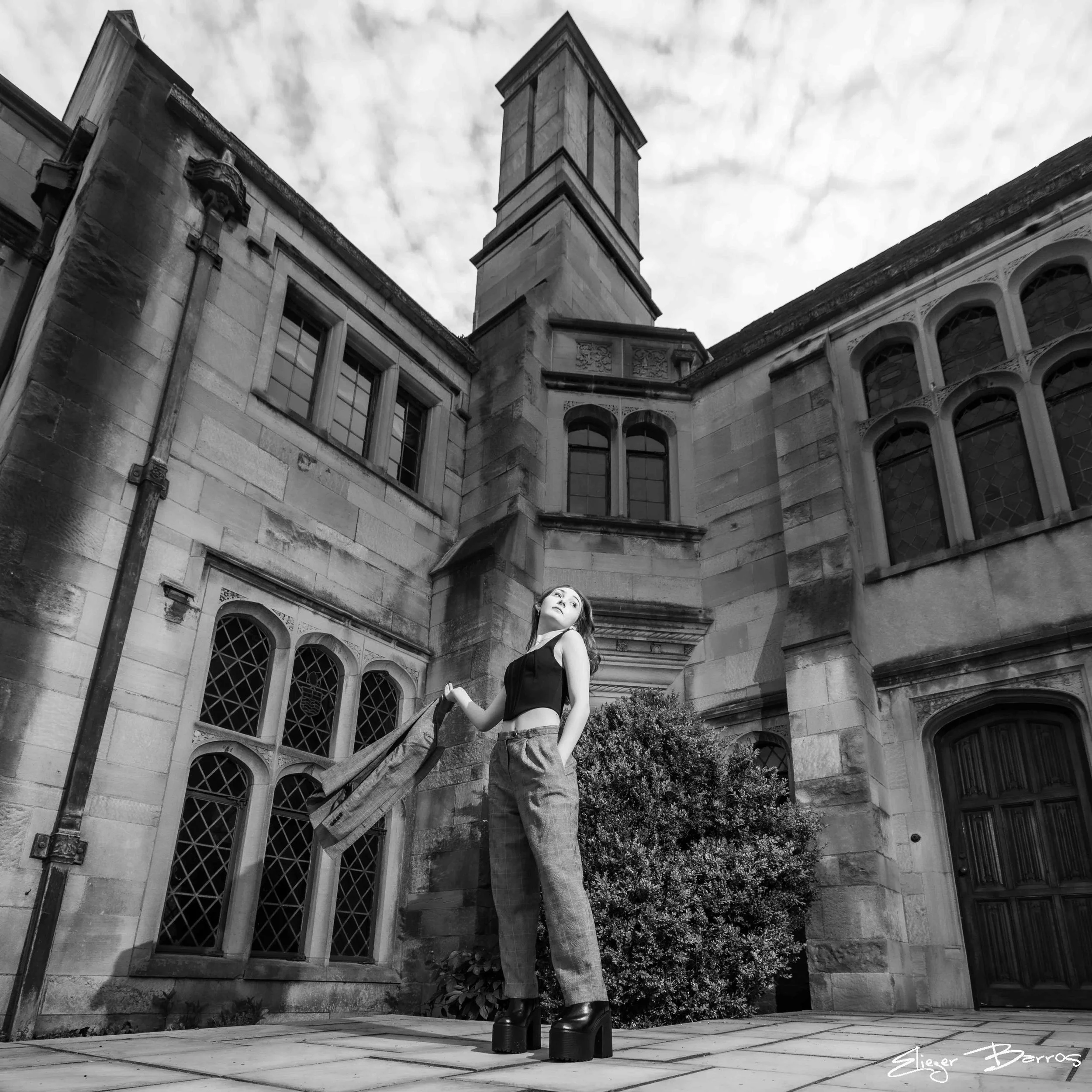 A woman with long hair standing in front of a historic stone building with a tower, multiple arched windows, and decorative stonework. She is wearing a sleeveless top, jeans, and platform shoes, posing with her hand on her hip and looking slightly up