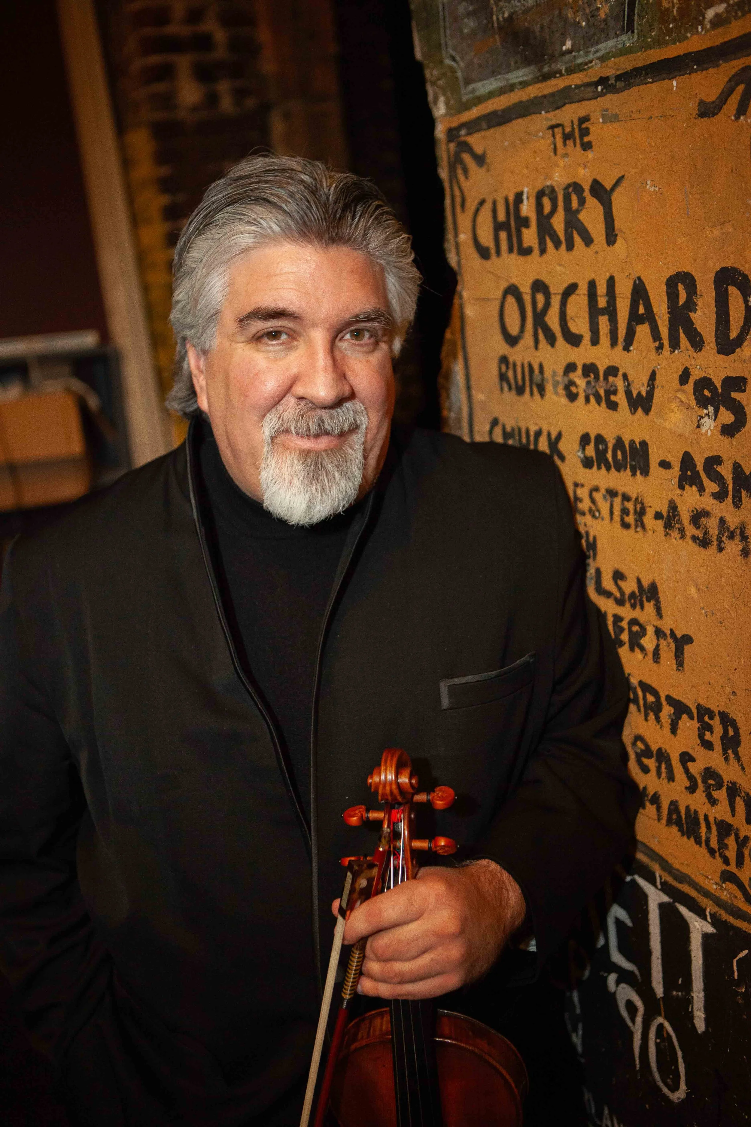 A man with gray hair and a beard playing a violin next to a brick wall with a painted sign that reads 'The Cherry Orchard Run & Crew' and other partially visible words.