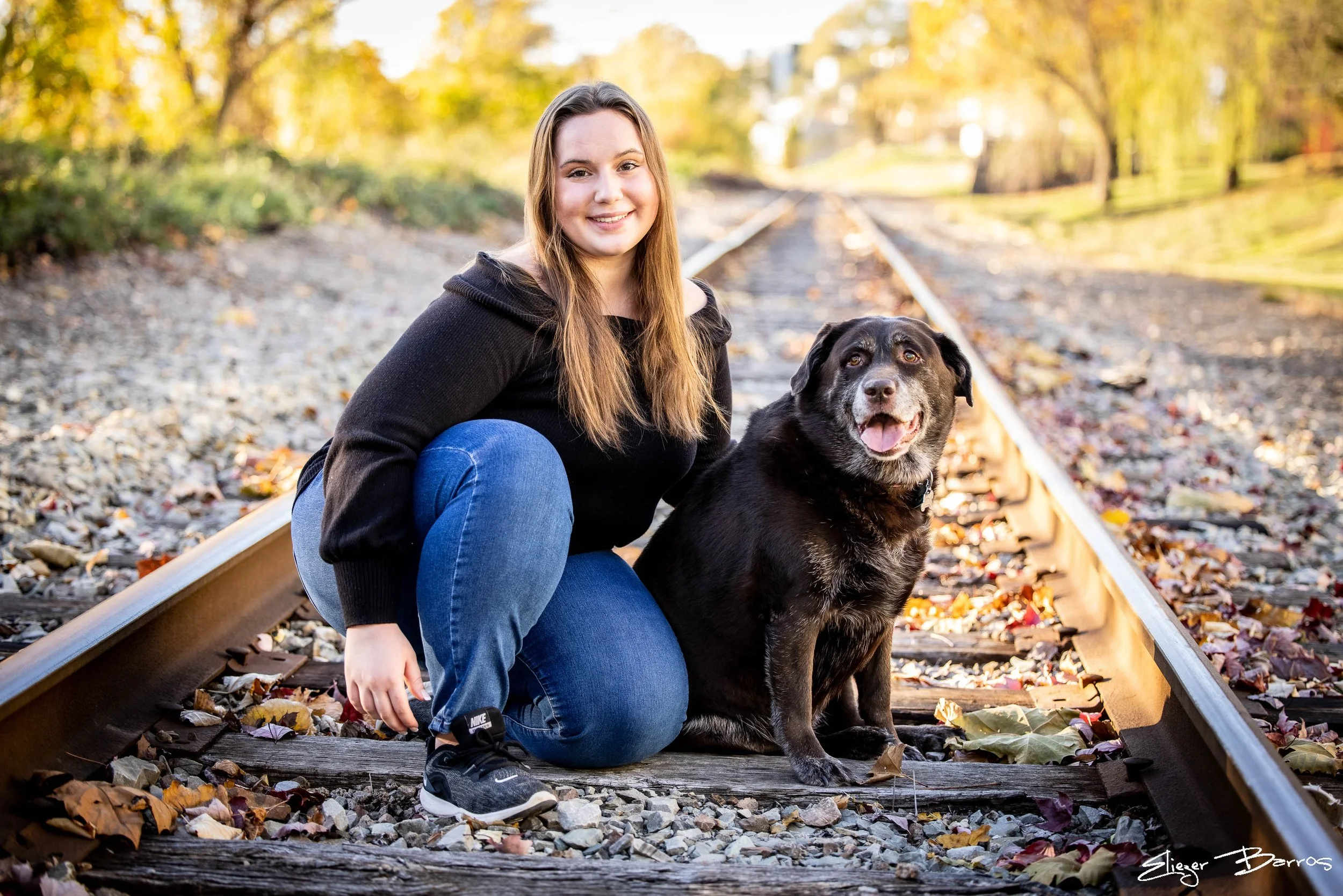 A young woman with long hair, wearing a black hoodie, kneeling on a railroad track with autumn leaves scattered around, smiling next to a black dog with a gray face in a park setting.