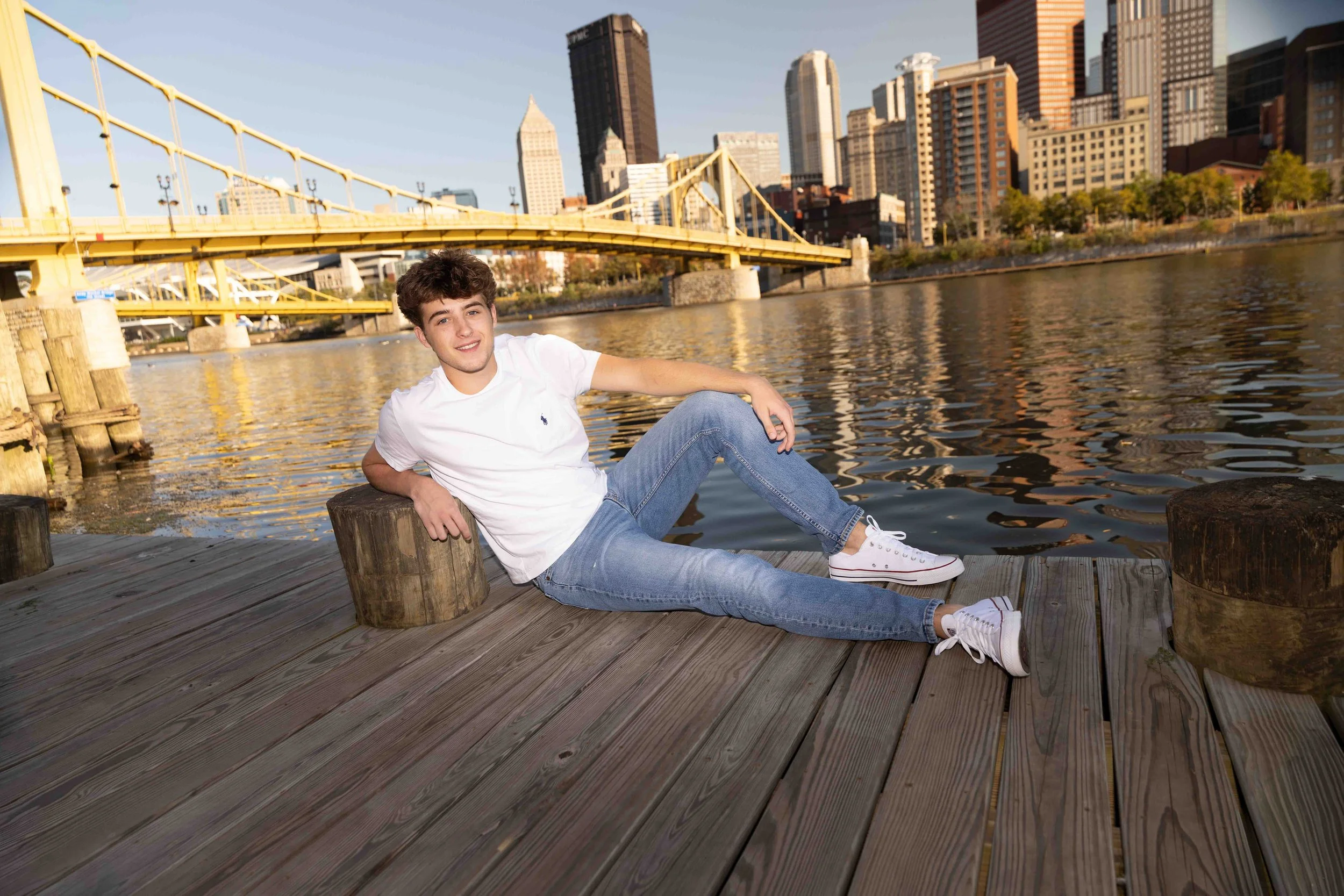 Young man sitting on a wooden dock by the water with city skyline and bridge in the background.