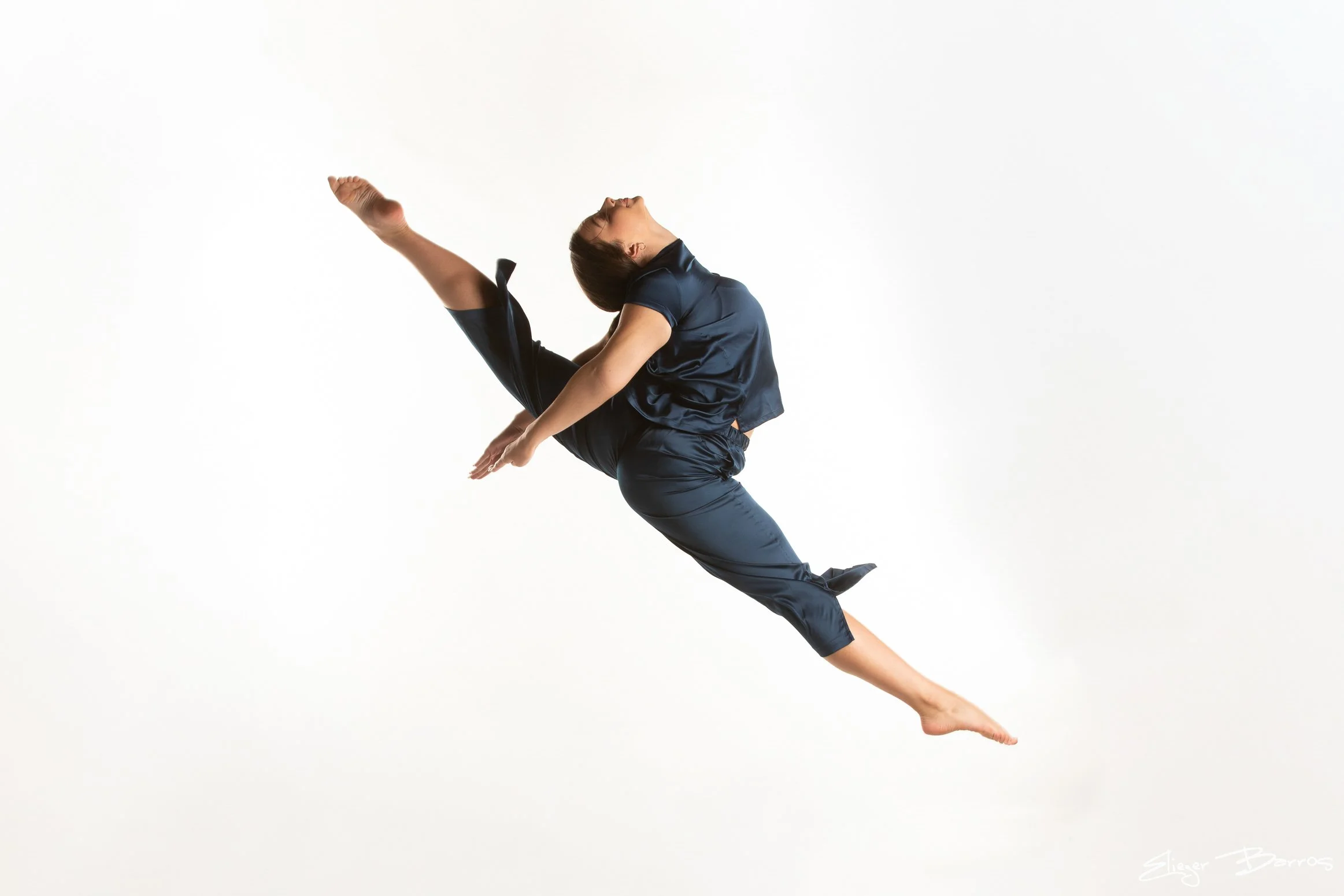 A woman in dark athletic clothing performing a high leg kick and stretching against a plain white backdrop.