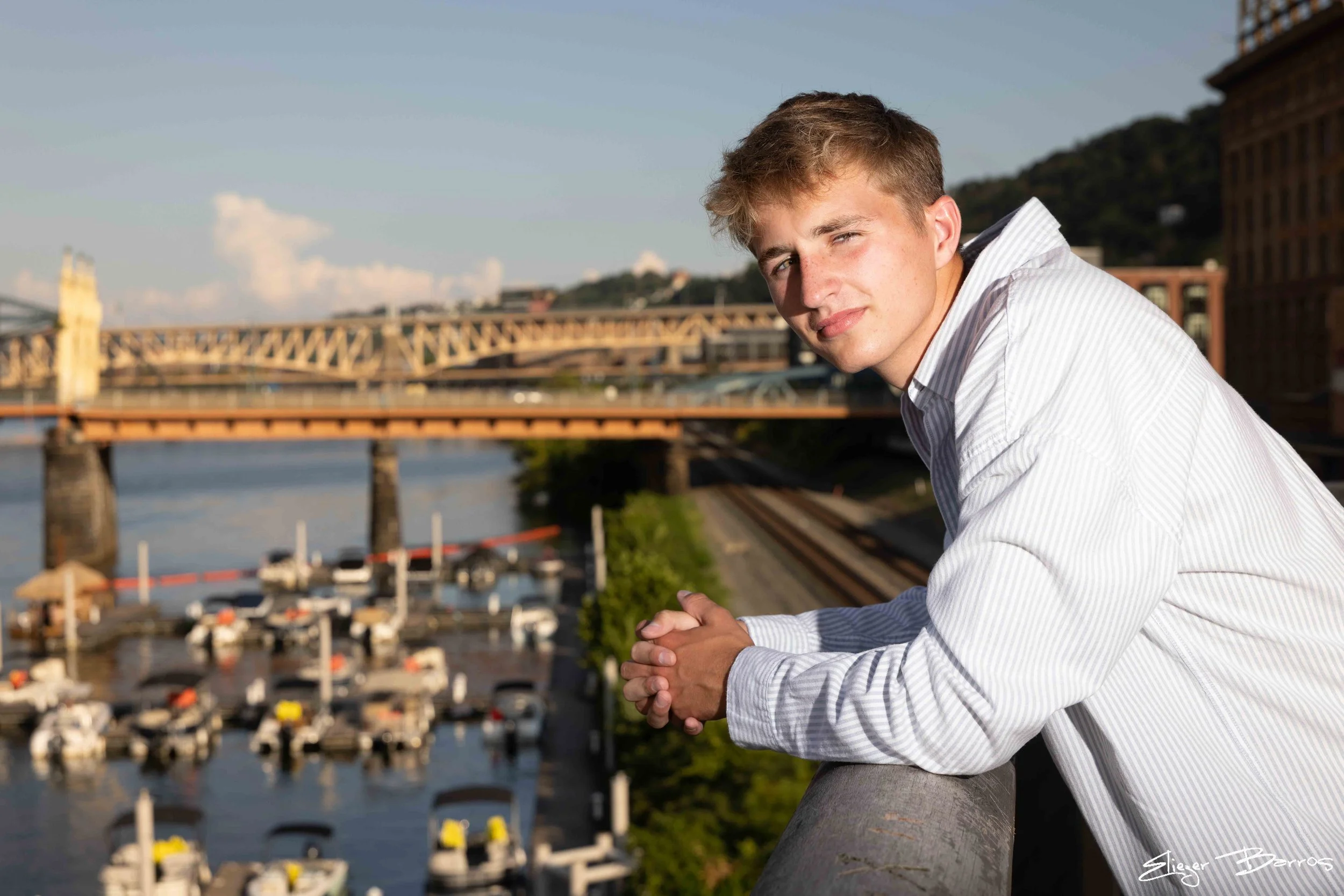 A young man with light brown hair and fair skin wearing a white striped shirt, sitting by a waterfront with city buildings and a bridge in the background.