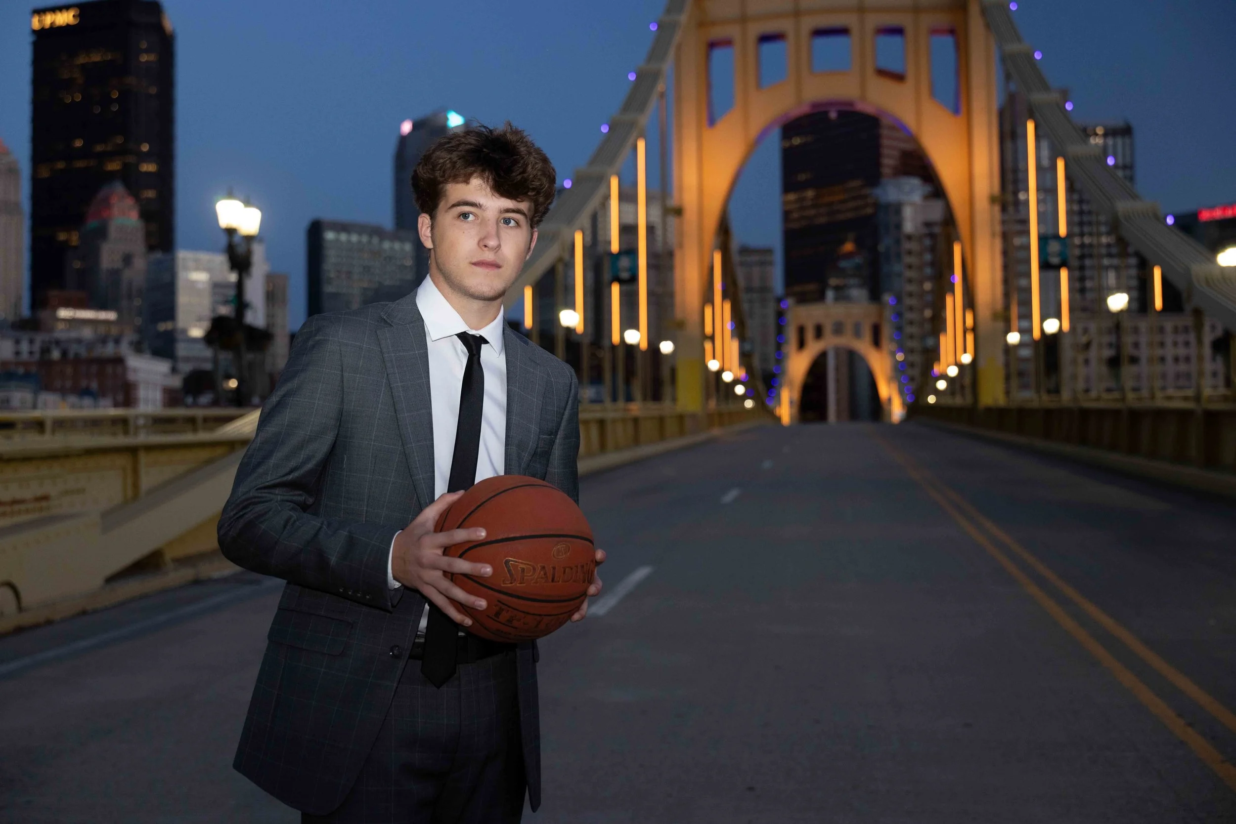 A young man in a suit holding a basketball on a city street at dusk with illuminated buildings and a bridge in the background.