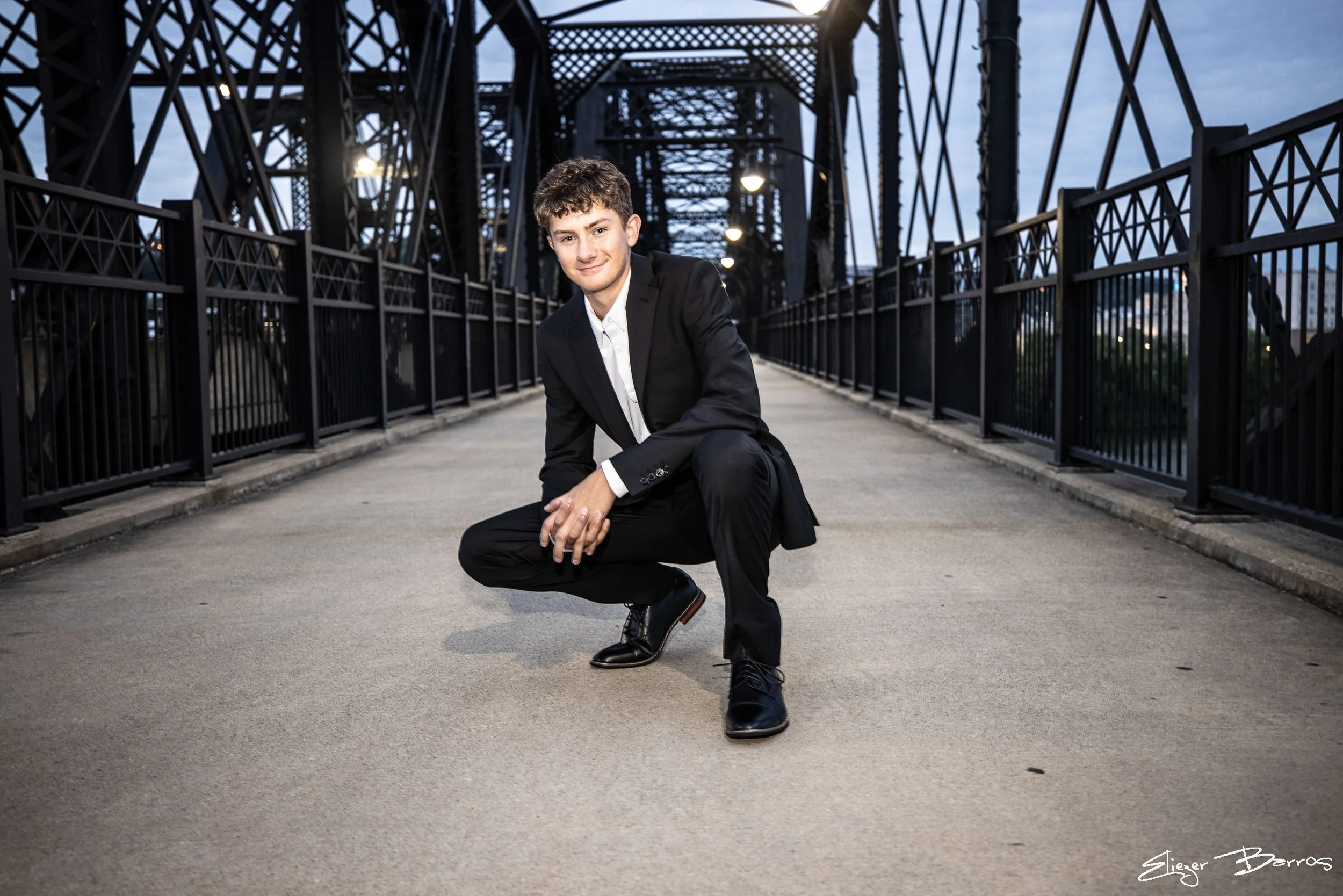 A young man in a black suit crouching on a bridge with metal framework, illuminated by streetlights, during twilight.
