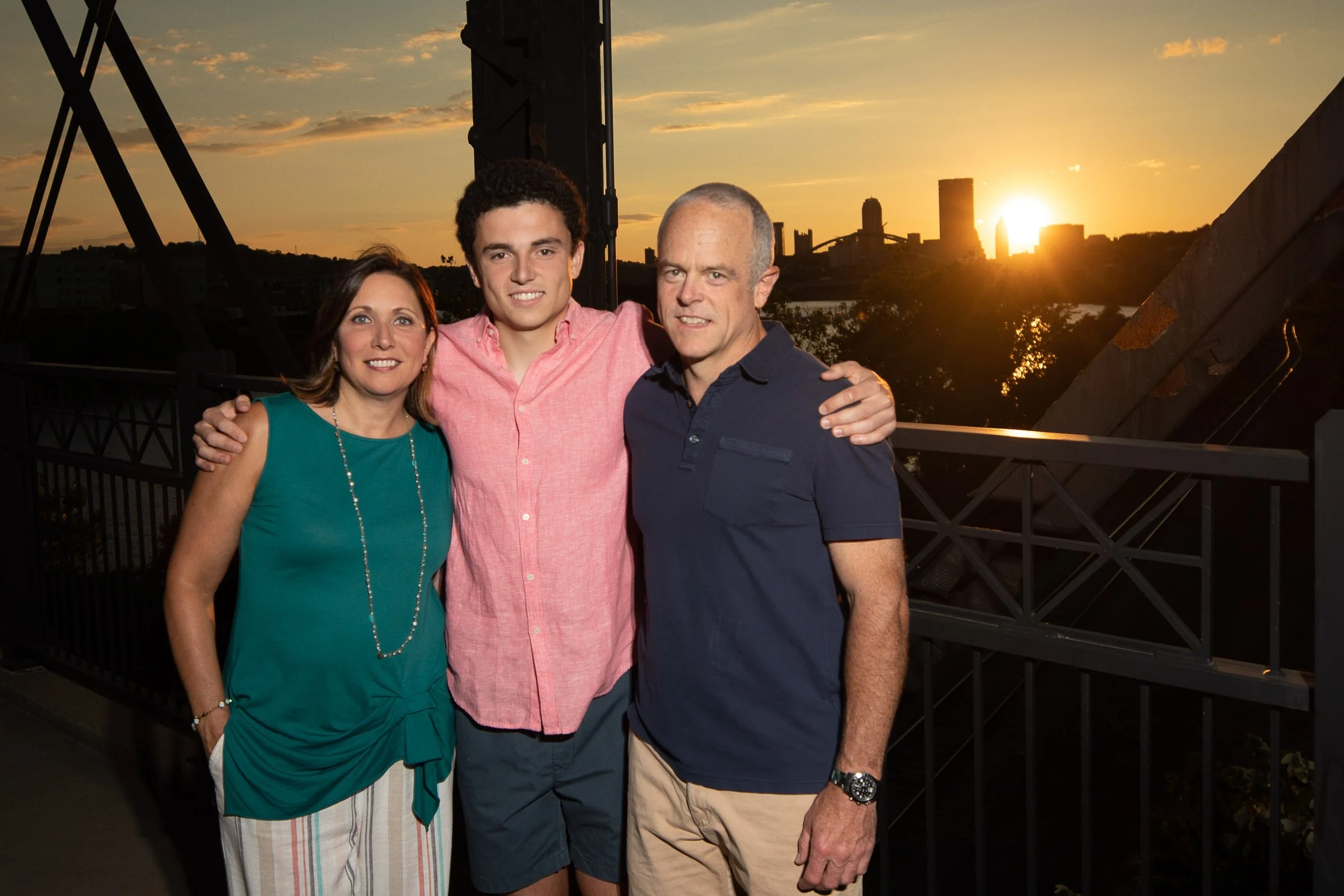 Three people, a woman and two men, standing together on a bridge at sunset with a city skyline in the background.