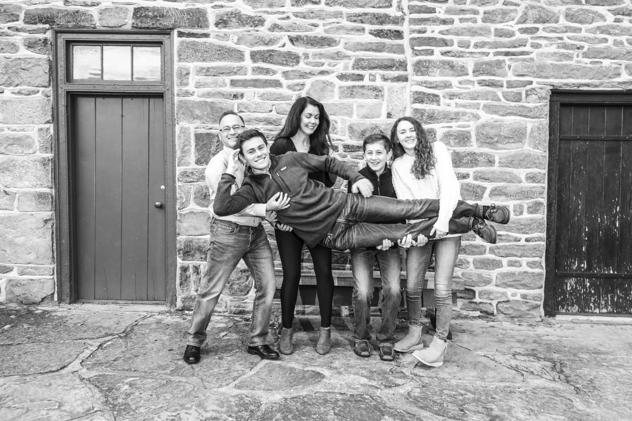 A group of six people, including three boys and three girls, standing against a stone wall. One boy is lying horizontally across the arms of the other five, all smiling and posing for the photo.