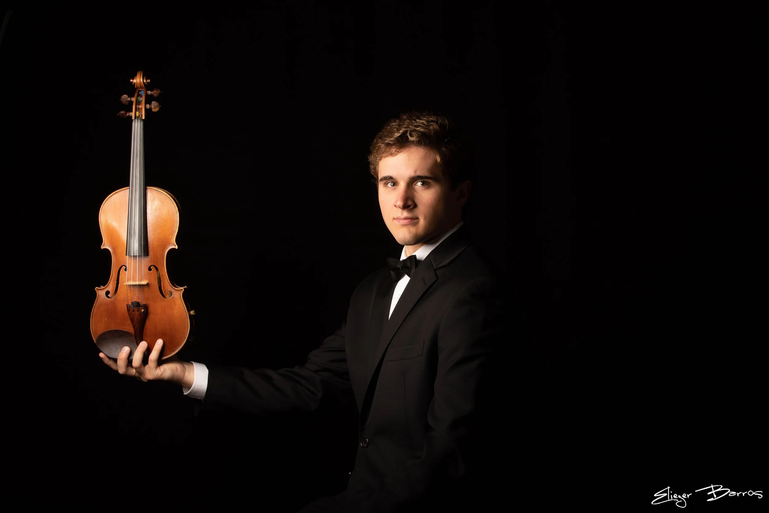 A young man in formal attire holding a violin against a black background.