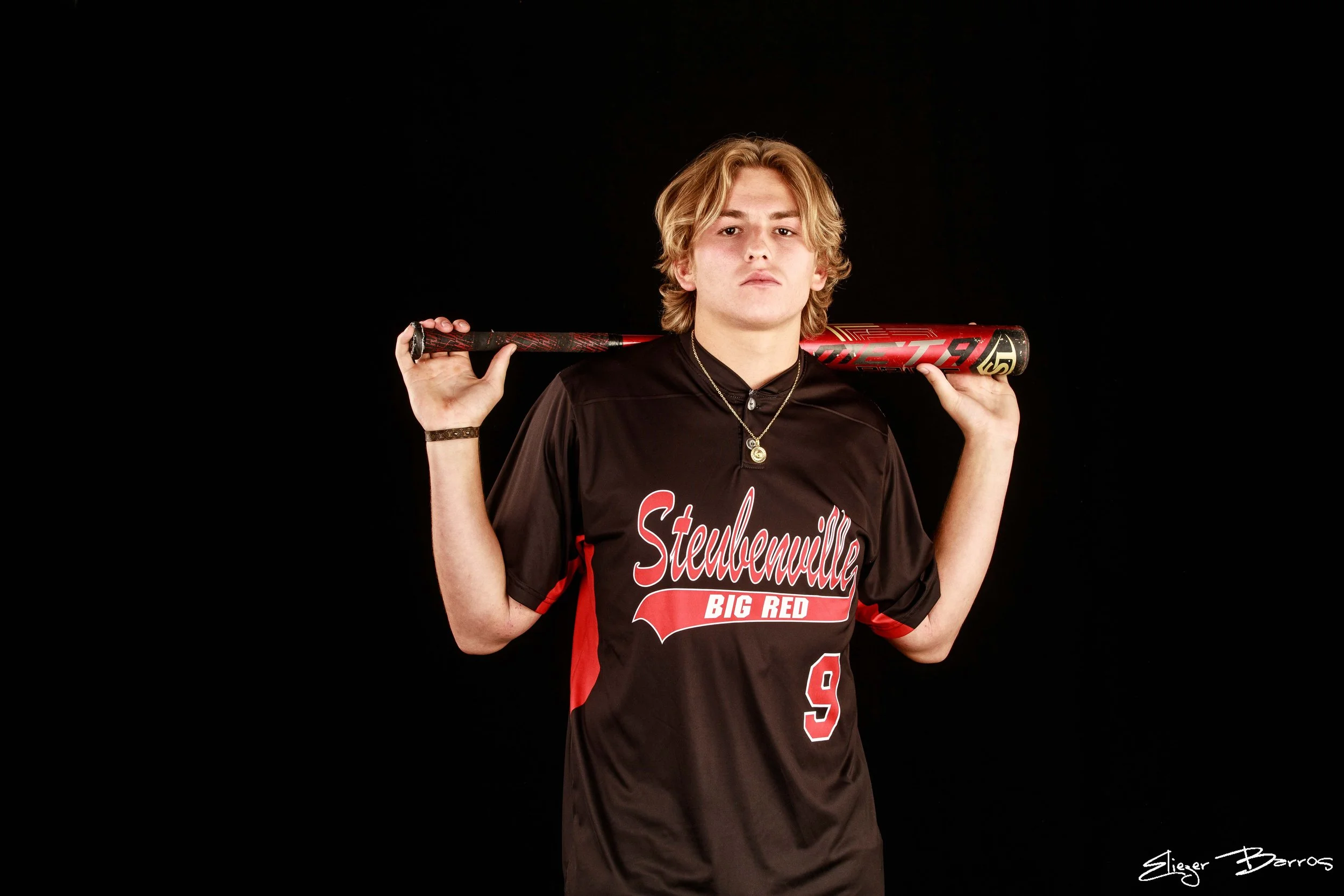 A young man with shoulder-length blonde hair holding a baseball bat over his shoulders, wearing a black and red sports jersey with the words 'Steulhemully Big Red' and the number 9, against a black background.