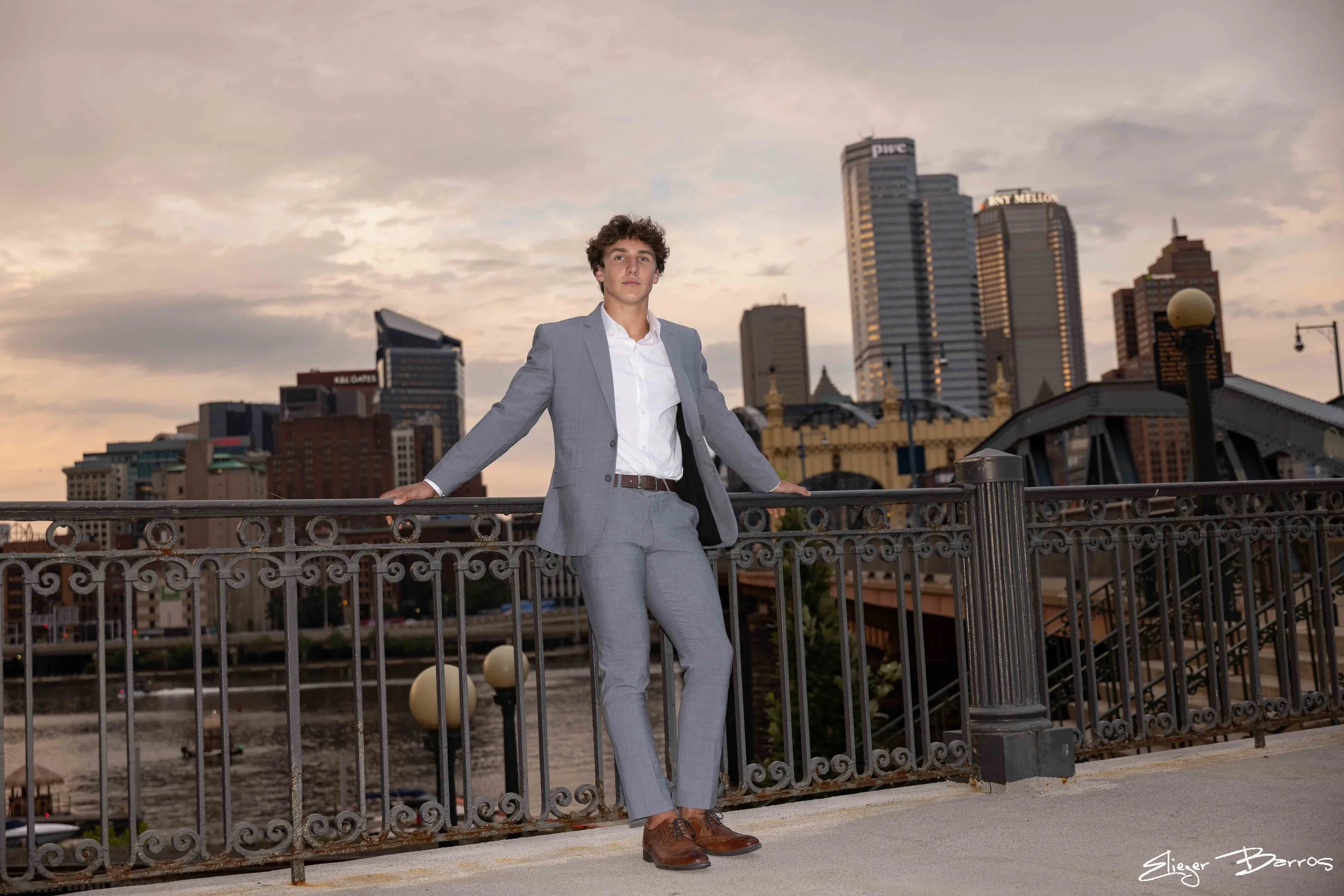 A young man in a gray suit posing on a bridge in an urban setting at sunset with tall buildings in the background.