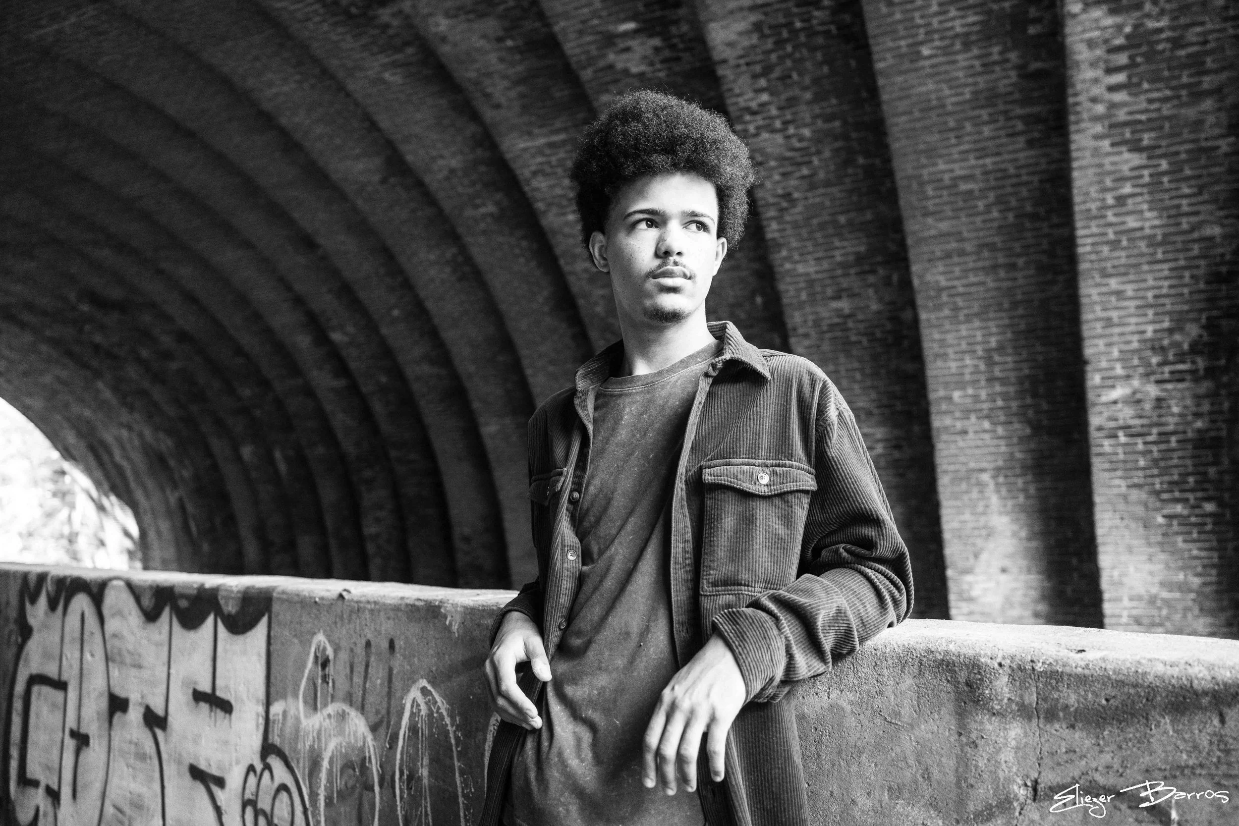 A young man with curly hair and a mustache stands under a curved brick bridge, looking off into the distance with a serious expression, wearing a denim jacket and layered clothing.