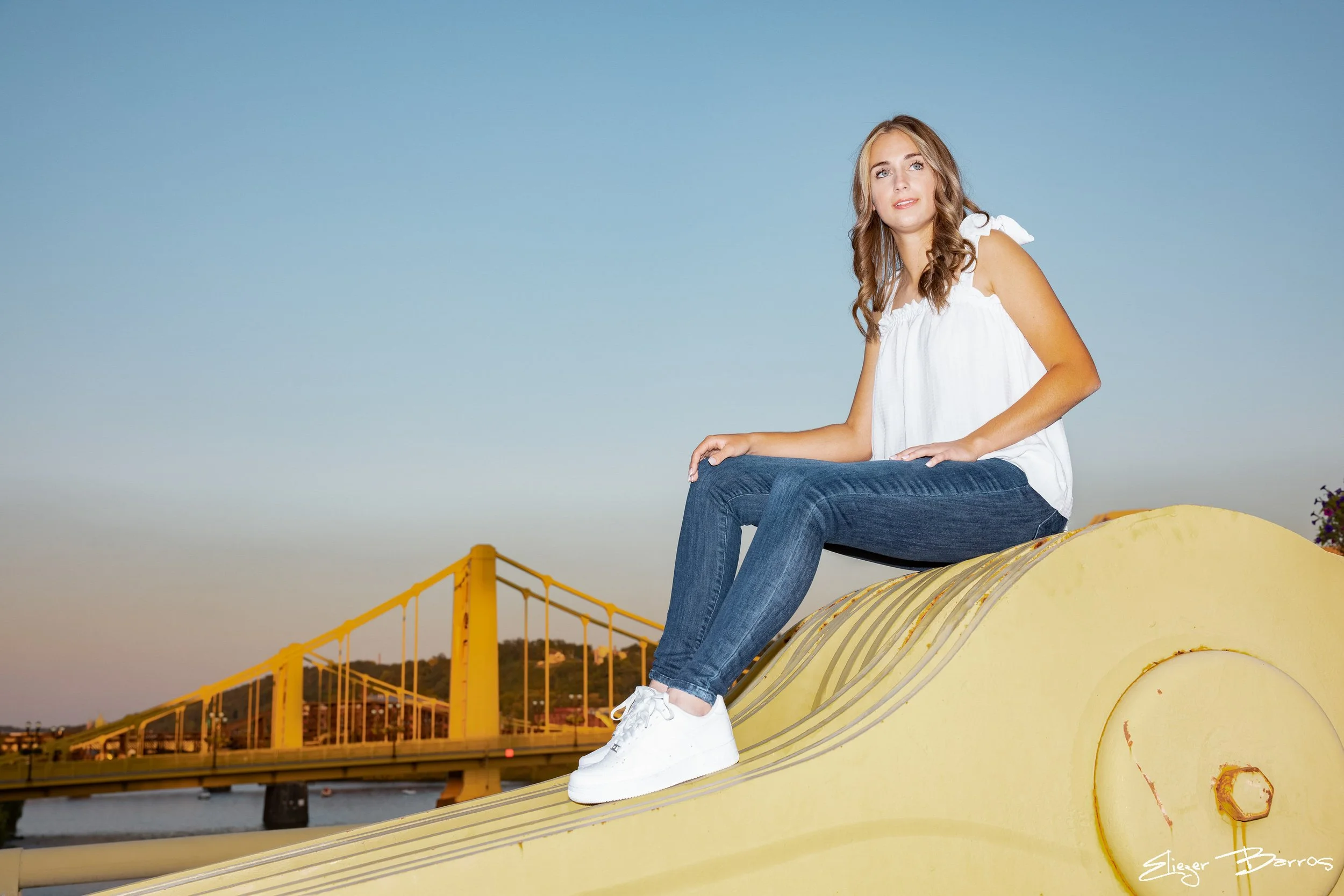 Senior Pictures Pittsburgh at Roberto Clemente Bridge. Young woman in white top and jeans sitting on the bridge during her outdoor urban senior photoshoot with Pittsburgh skyline behind her.