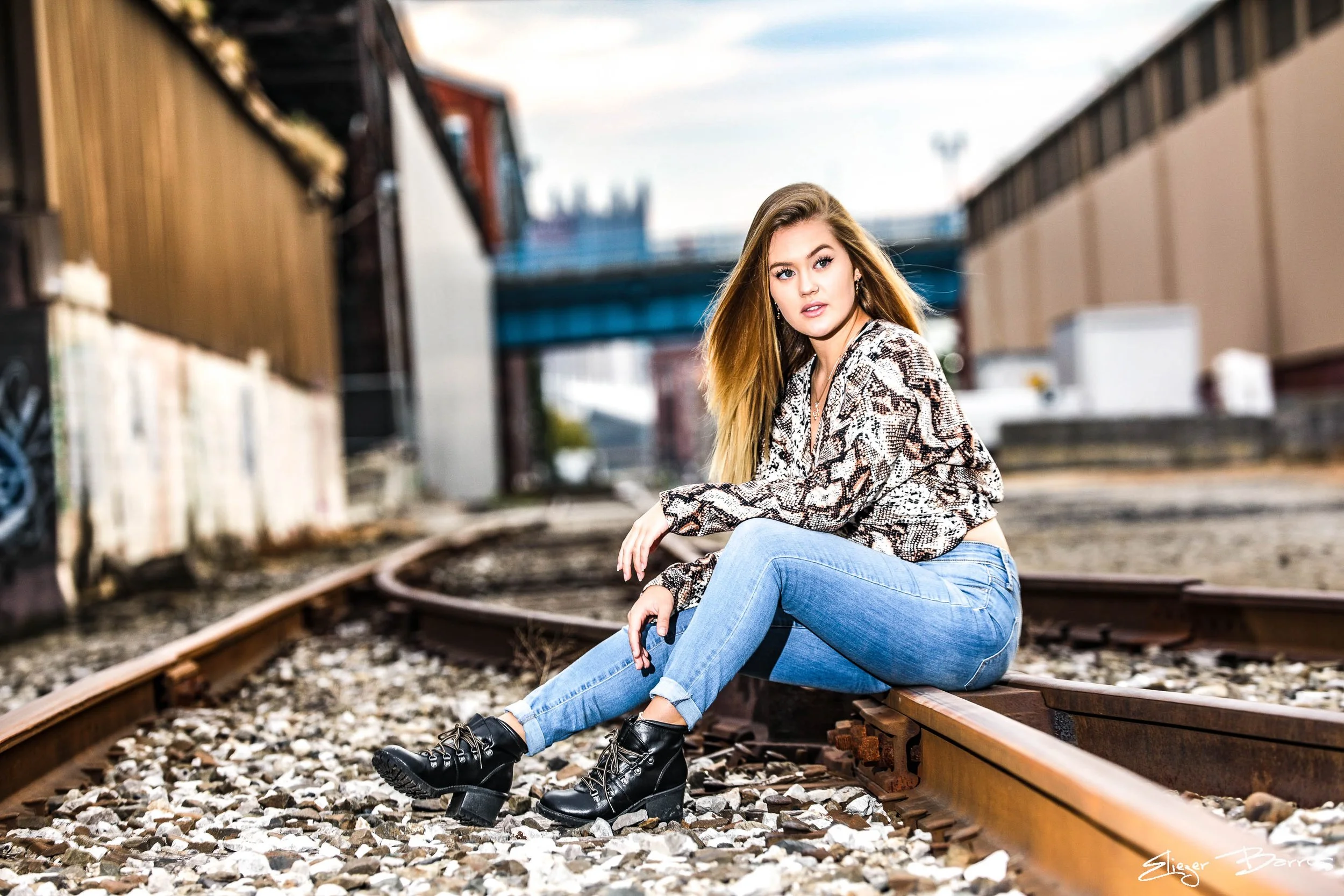 A young woman sitting on train tracks outdoors, wearing a snakeskin patterned jacket, blue jeans, and black boots, with a blurred urban background.