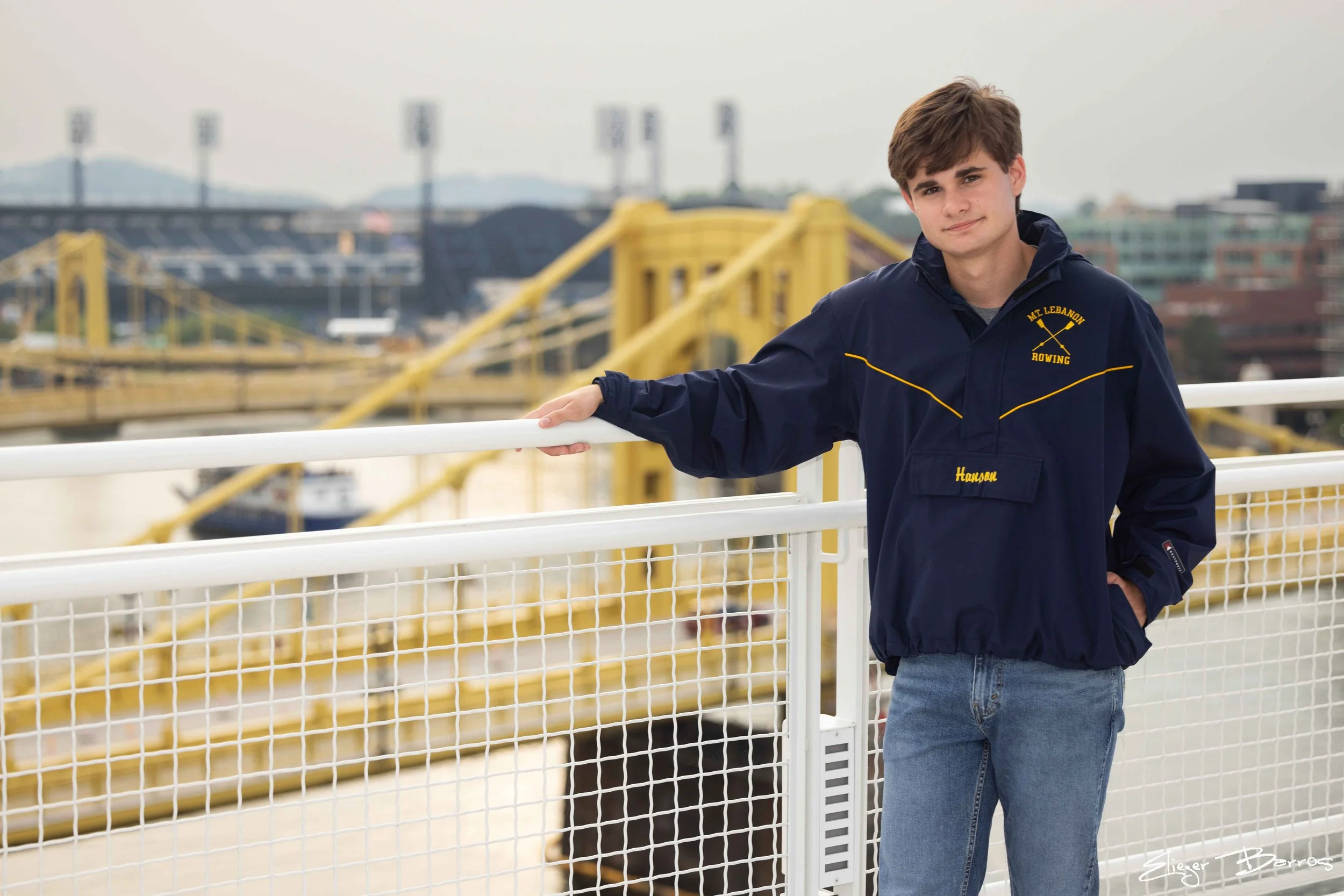 A teenage boy in a navy blue jacket with yellow embroidery that says "Mt. Lebanon Rowing" and his name "Hanson" in yellow, standing on a boat dock, with a yellow bridge, water, and city buildings in the background.