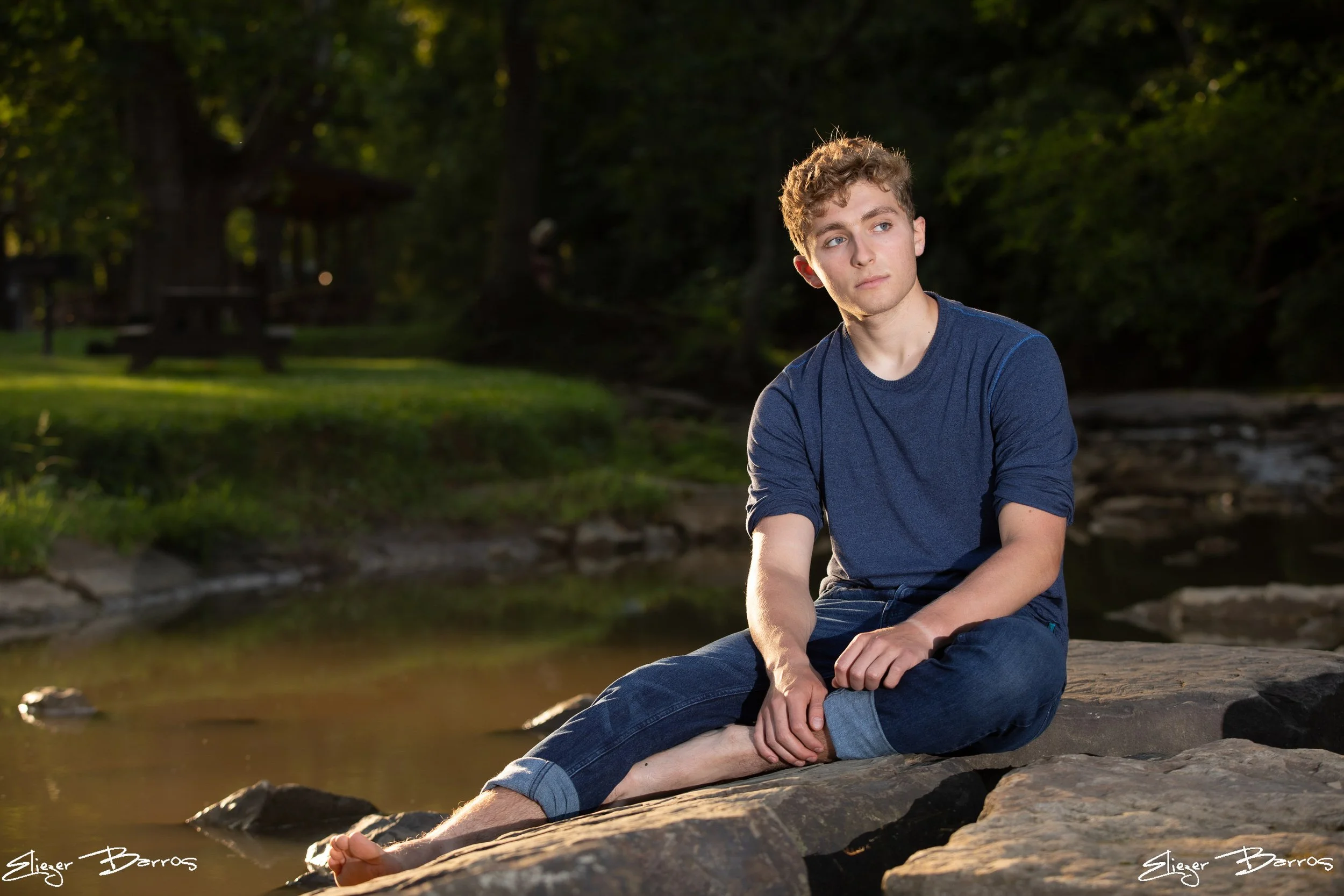 Senior Pictures Pittsburgh at Mingo Creek Park. Young man in a blue t-shirt and jeans sitting on rocks by the water at the scenic Ebenezer Covered Bridge for relaxed outdoor senior pictures.