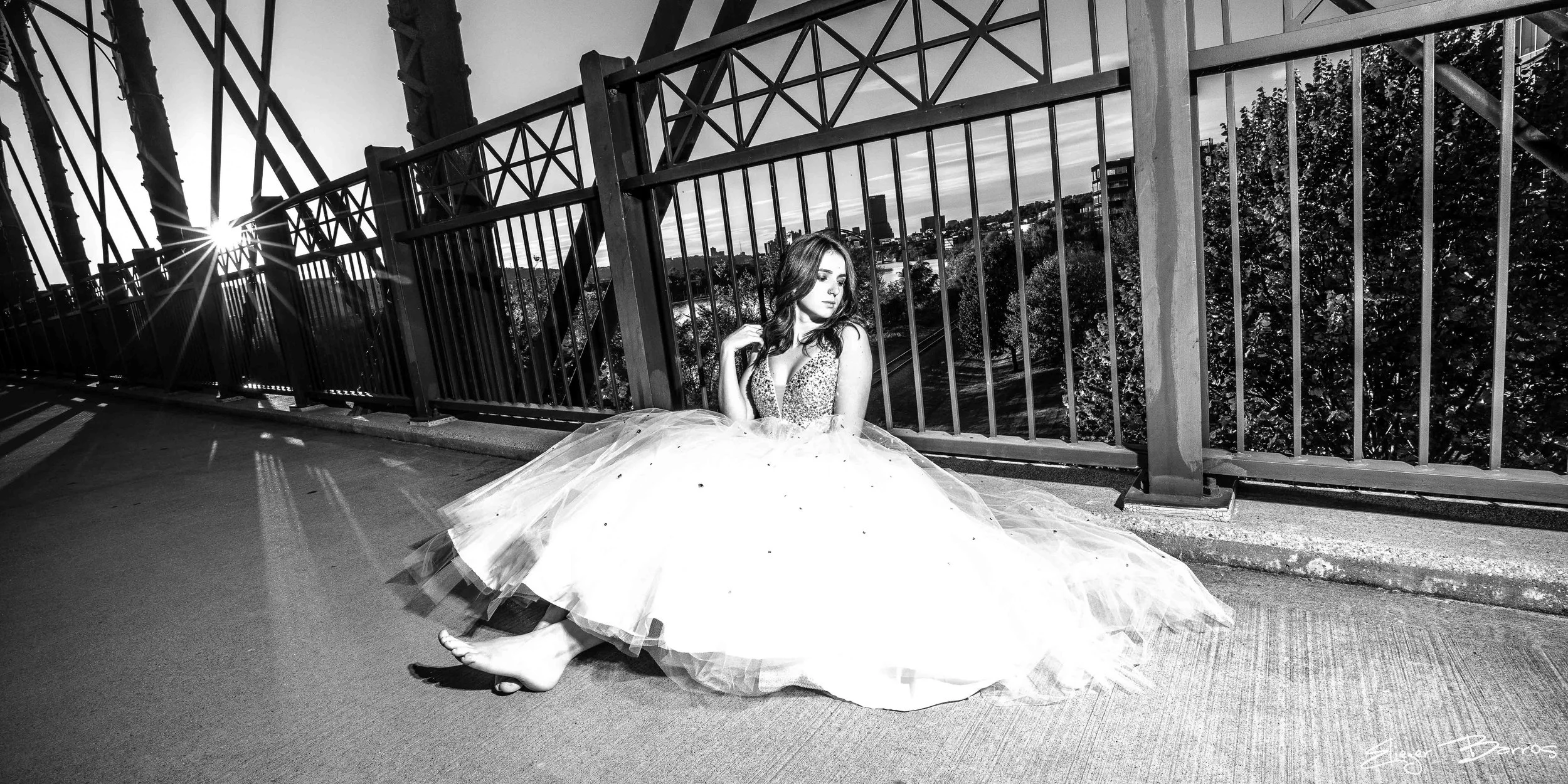 A woman in a ball gown sitting on a sidewalk near a metal fence with the cityscape in the background, during sunset, in black and white.