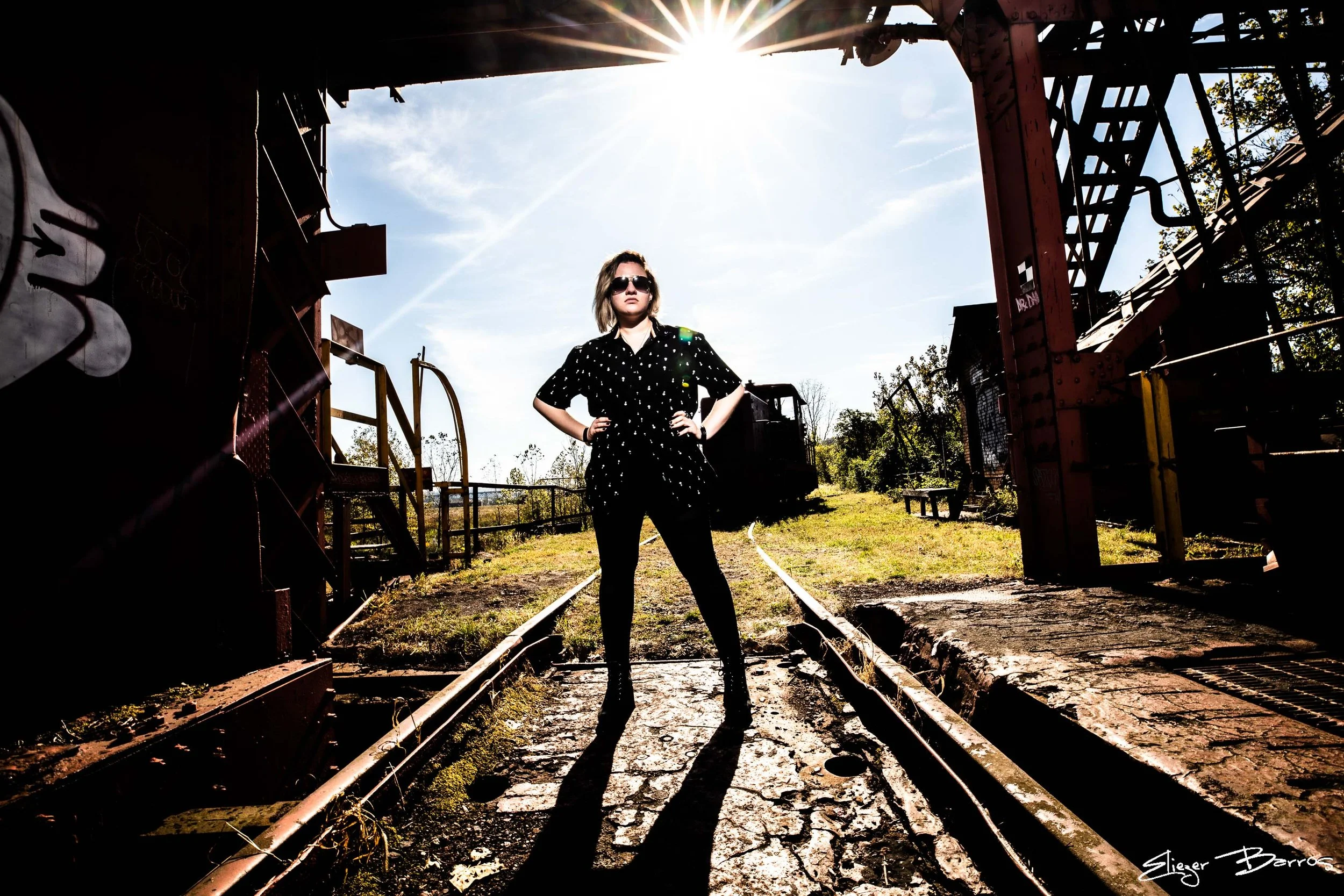 Senior Pictures Pittsburgh at Carrie Furnace. Young woman in black outfit posing on railroad tracks with dramatic sun flare and industrial ruins for powerful outdoor senior pictures.