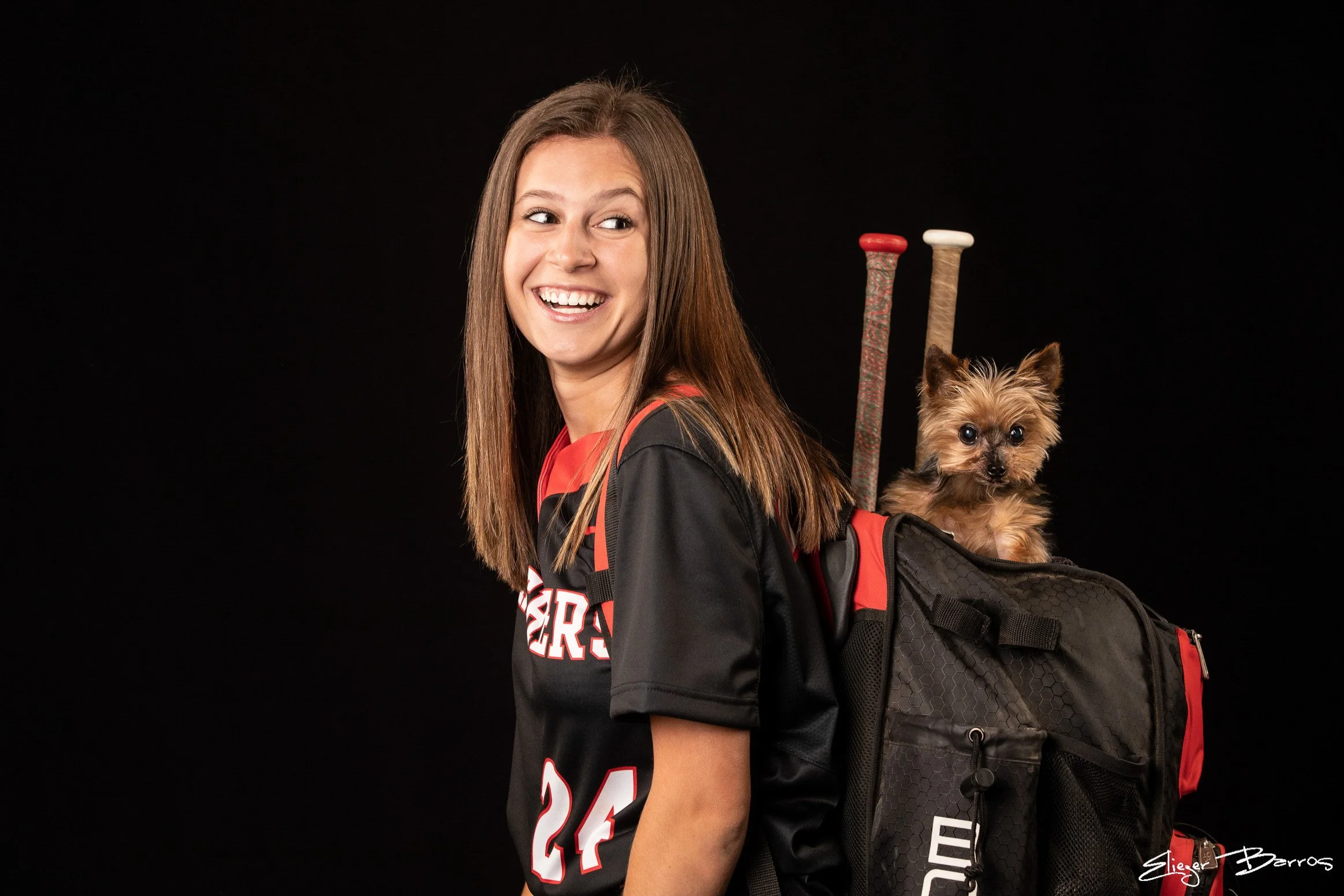 A young woman with long brown hair, wearing a sports jersey, carrying a black backpack with red accents, and a small dog in the backpack.