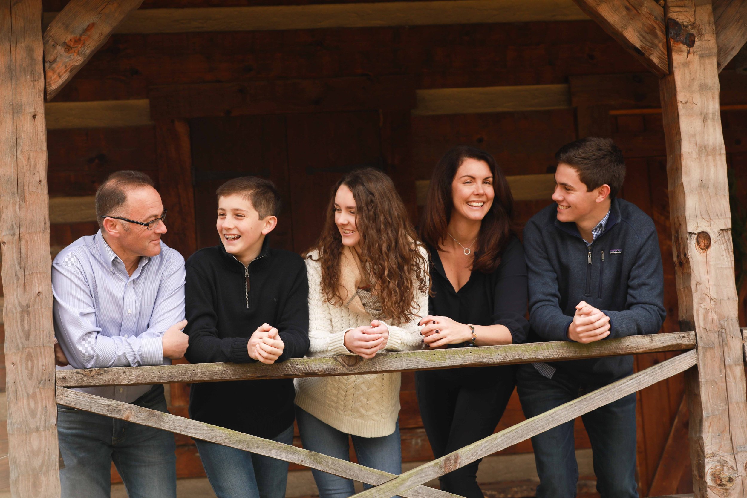 Group of six people, three adults and three teenagers, smiling and talking on a wooden balcony with a rustic wooden structure, possibly in a rural or outdoor setting.
