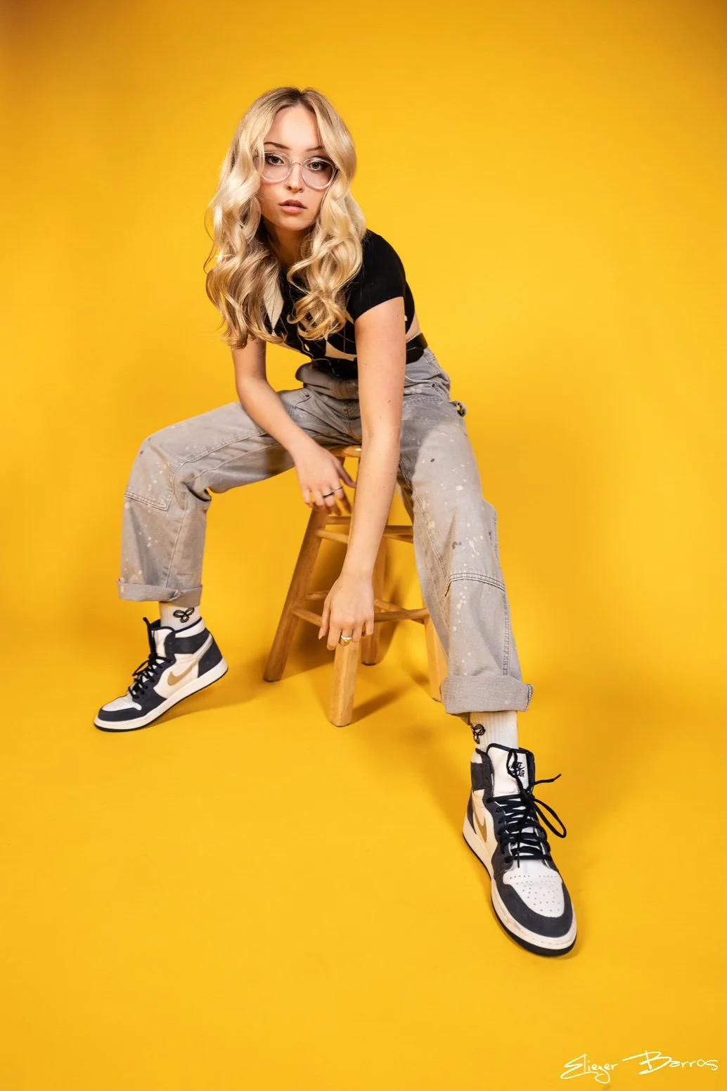 A young woman with blonde, wavy hair, wearing glasses, a black crop top, baggy gray pants, white socks, and black-and-white Nike sneakers, sitting on a small wooden stool against a bright yellow background.