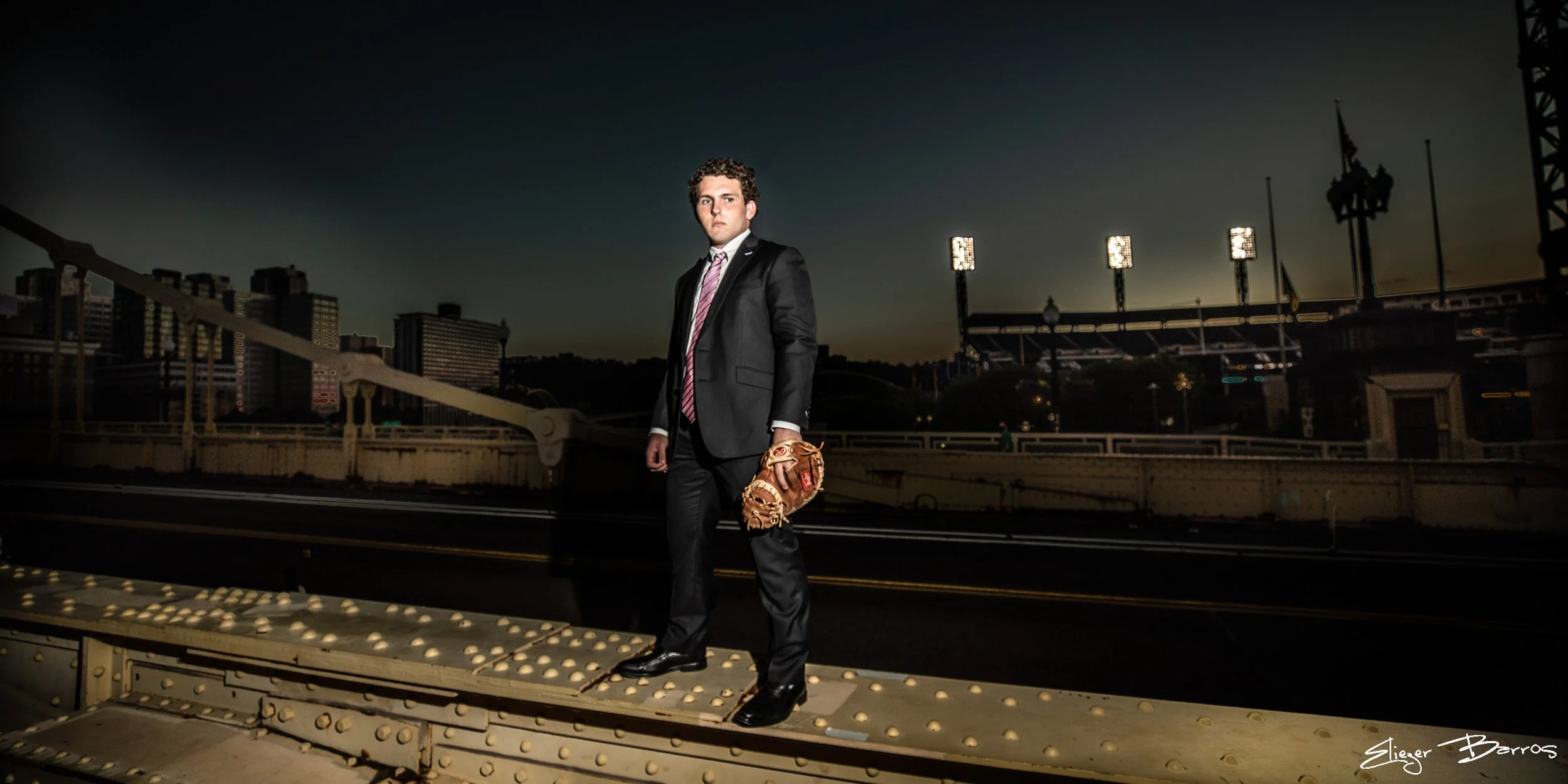 A young man in a dark suit with a pink striped tie stands on a bridge at dusk, holding a baseball glove in one hand, with city buildings and a stadium in the background.