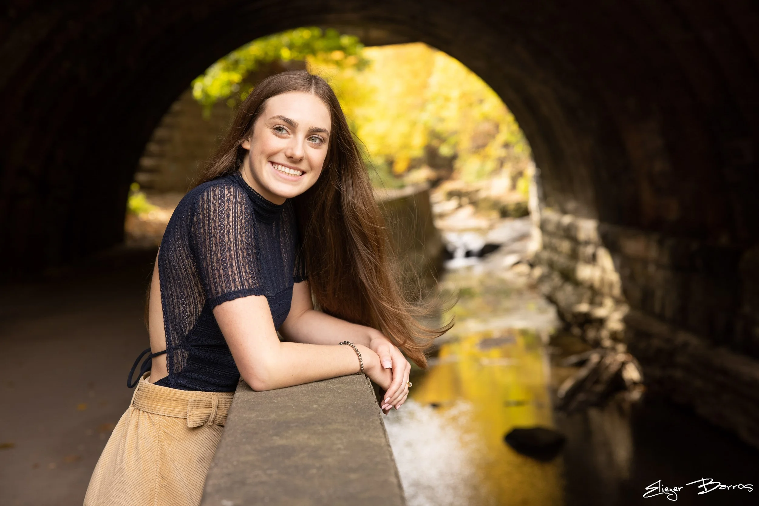 A young woman with long brown hair and a black lace top smiling while leaning on a stone railing near a small creek under a stone bridge with fall foliage in the background.