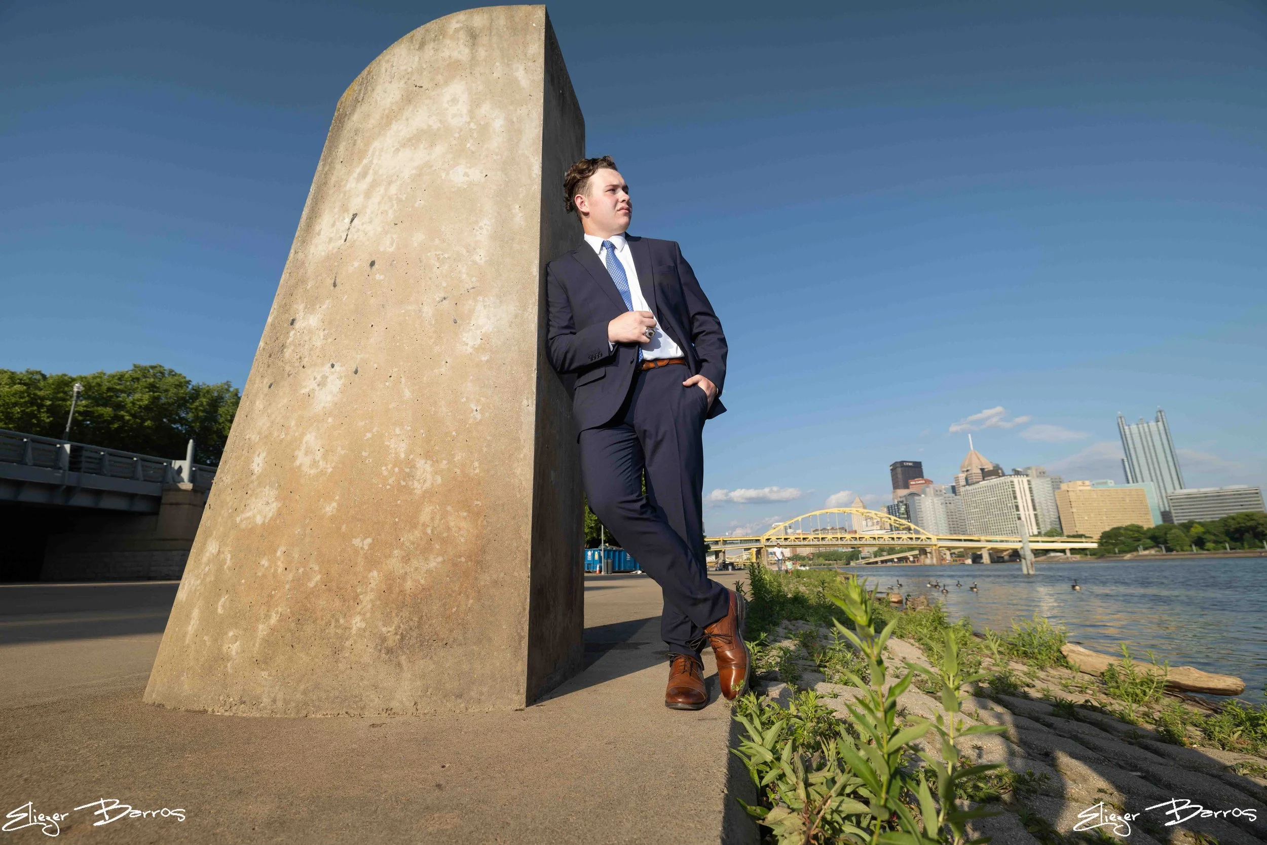 A man in a business suit leaning against a large concrete sculpture by a river with a city skyline in the background.