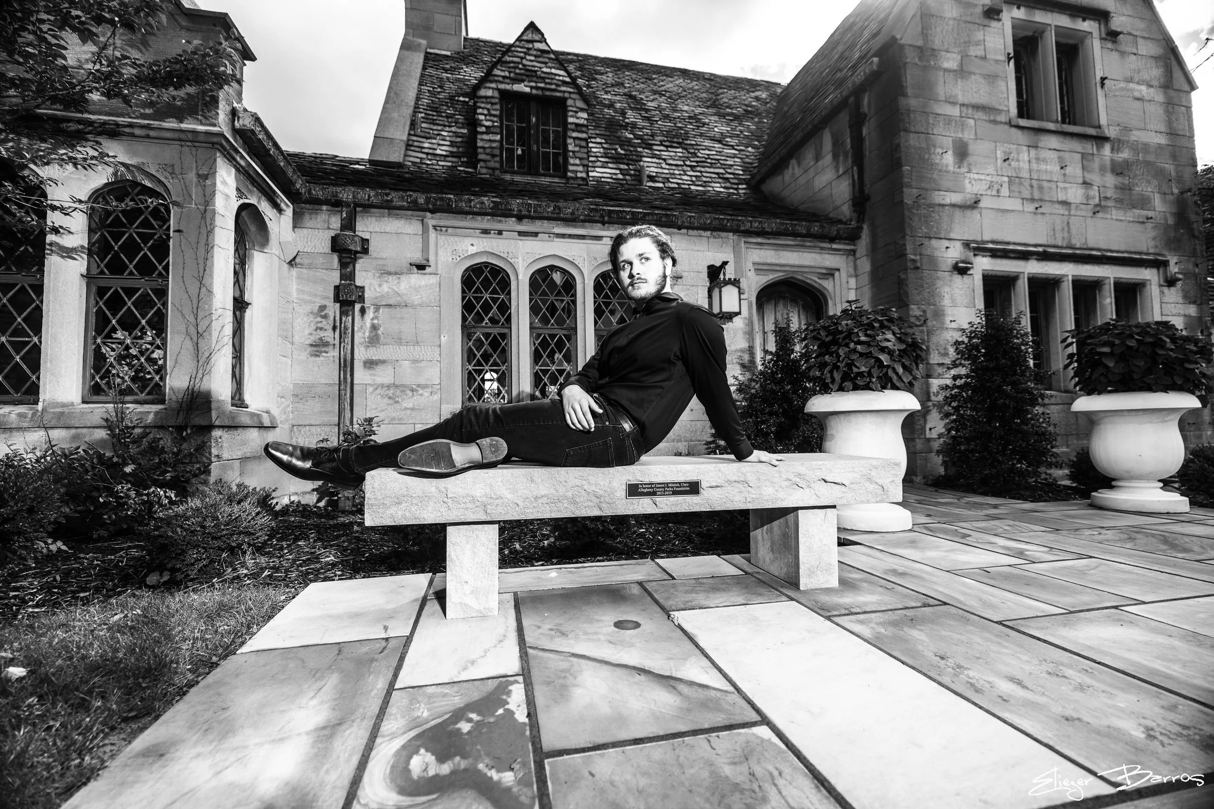 Black and white photo of a young man with beard and dark hair, sitting on a stone bench in front of a historic stone building with arched windows, surrounded by potted plants and shrubbery.