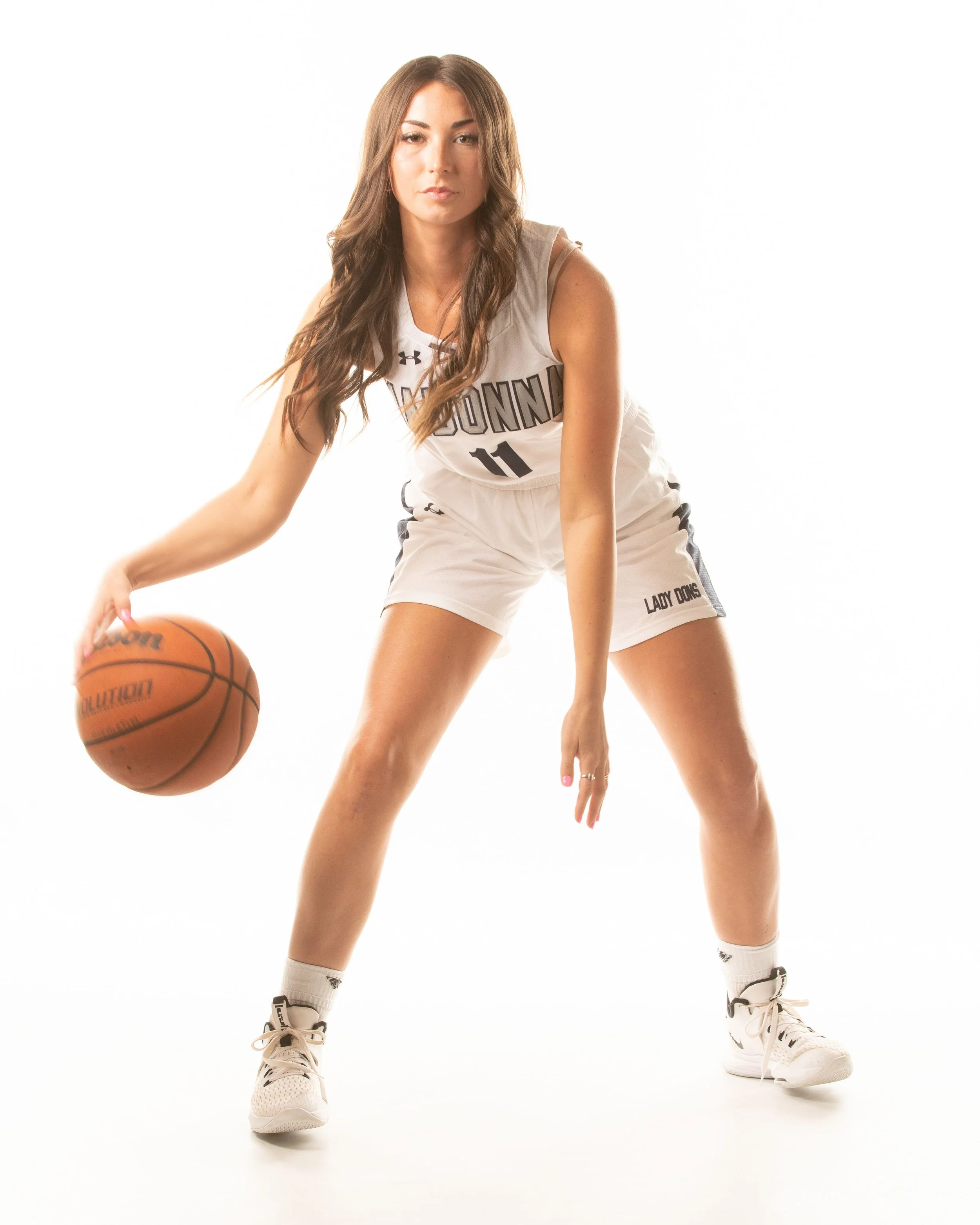 A young woman wearing a white basketball uniform with the number 11, white socks, and sneakers, dribbling a basketball in a pose against a plain white background.