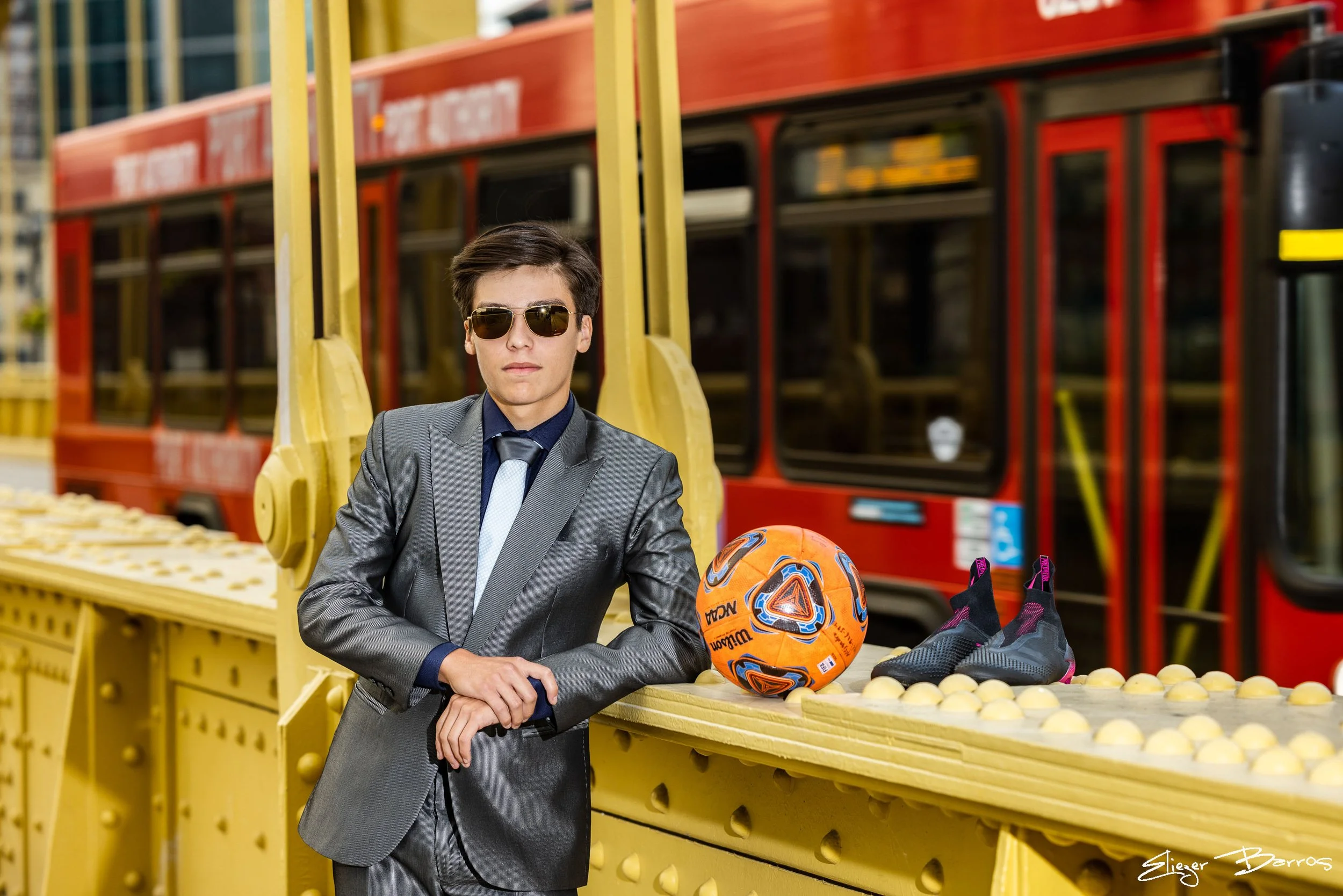 A young man in a gray suit and sunglasses leaning on a yellow banana display at a store, with a soccer ball and a pair of sneakers nearby.