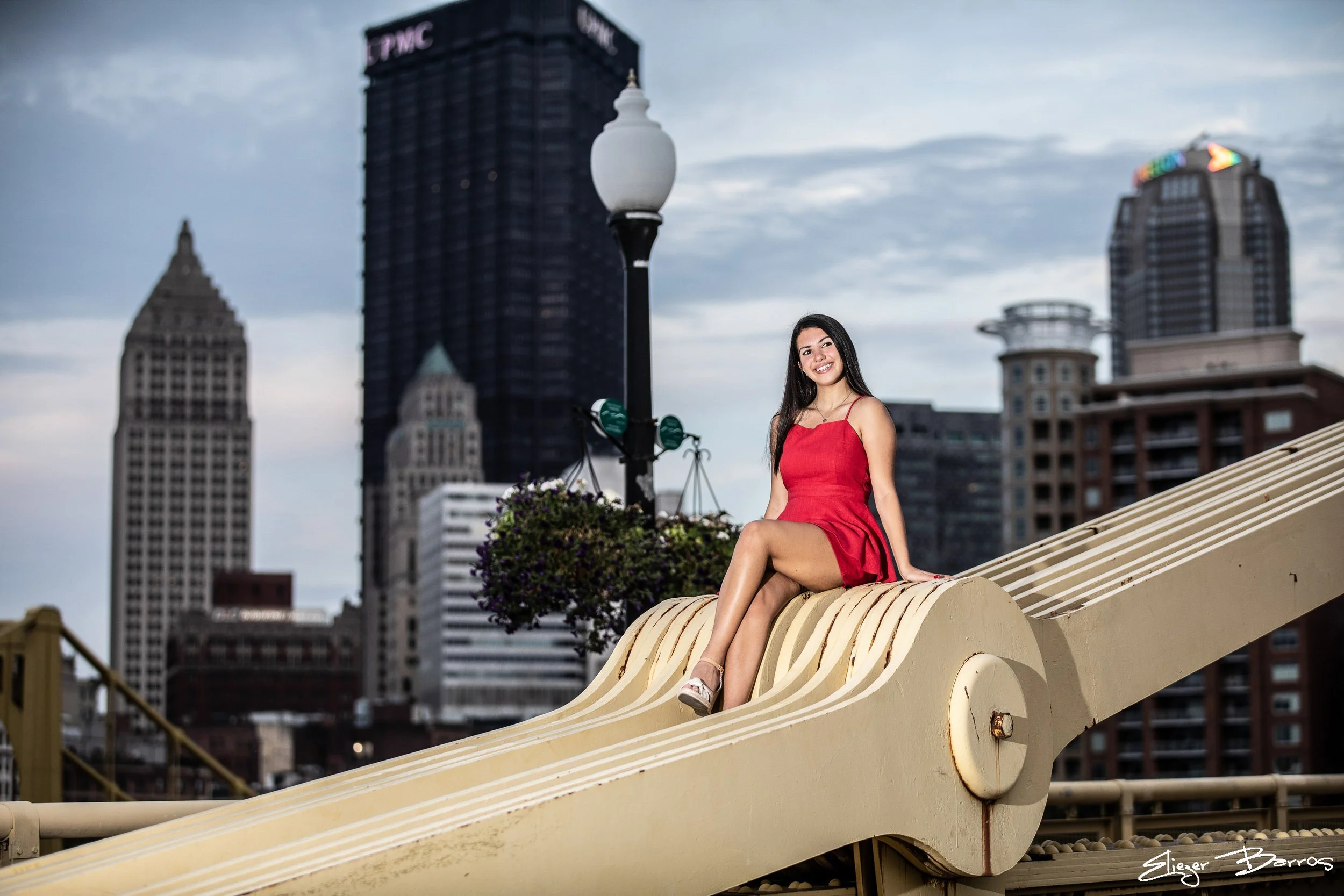 A woman in a red dress sitting on a large curved metal structure on a bridge with city skyscrapers in the background and a cloudy sky.