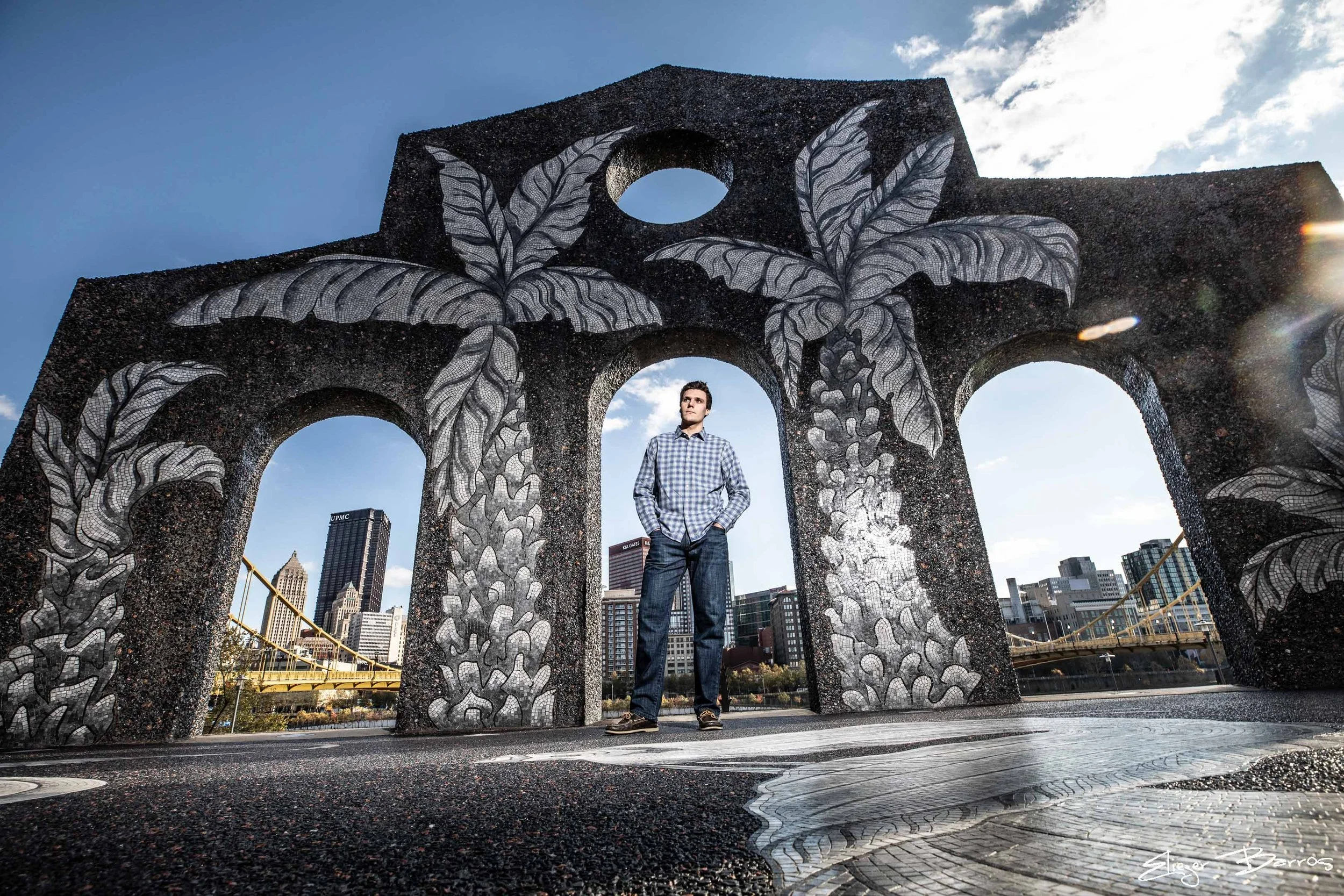 A man stands in front of a large decorative structure with black and white mosaic leaf designs and circular openings, with a city skyline in the background under a partly cloudy sky.