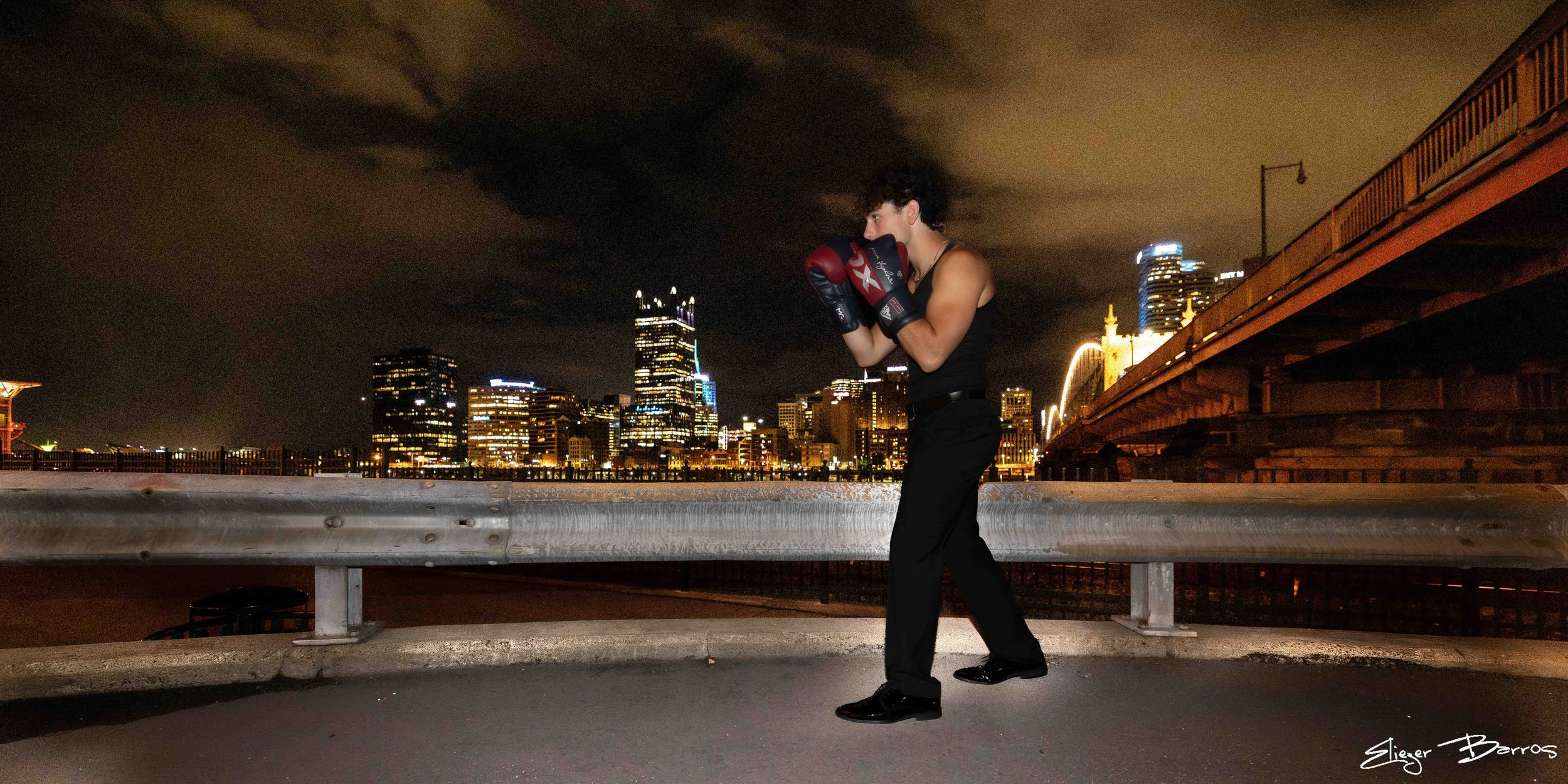 A woman in boxing gloves practicing boxing on a bridge at night with a city skyline in the background.