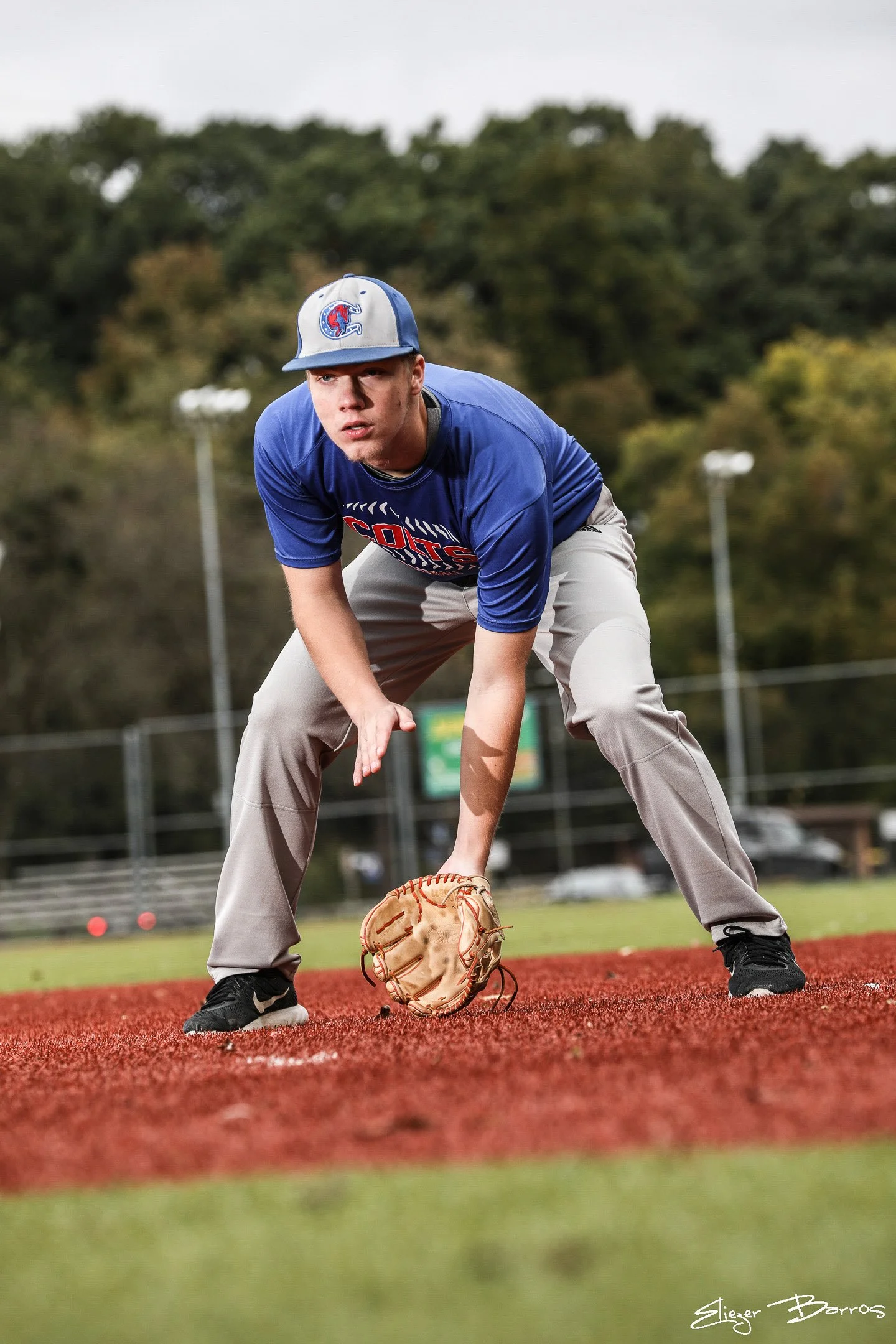 A young man in a blue sports jersey and gray pants is playing baseball on a field. He is wearing a gray and blue cap with a logo and is in a ready stance, holding a baseball glove with one hand while looking towards the pitcher. The background shows 