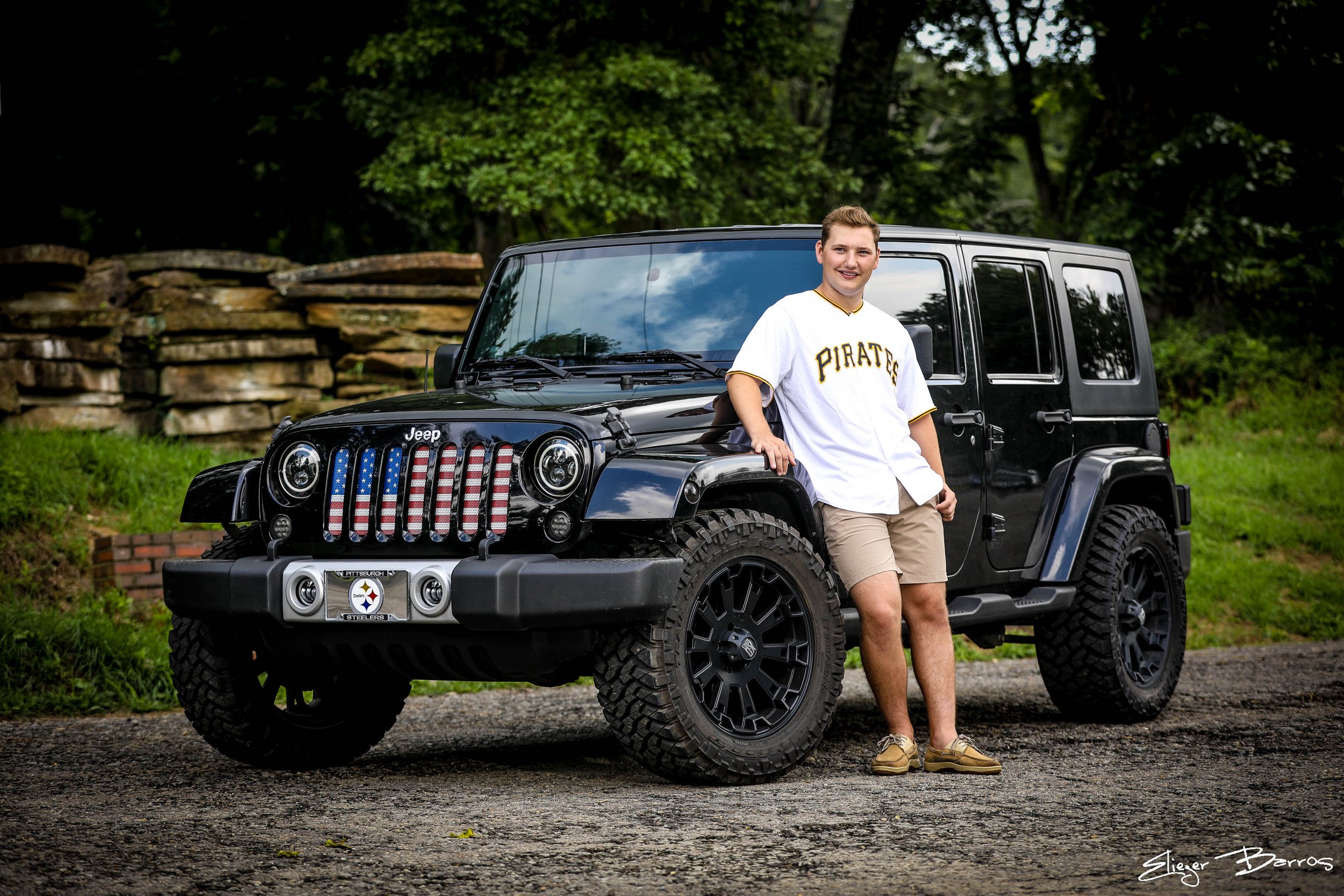A young man wearing a white Pittsburgh Pirates baseball jersey and khaki shorts standing beside a black Jeep Wrangler with an American flag-themed grille in an outdoor setting with green trees and a stone wall in the background.