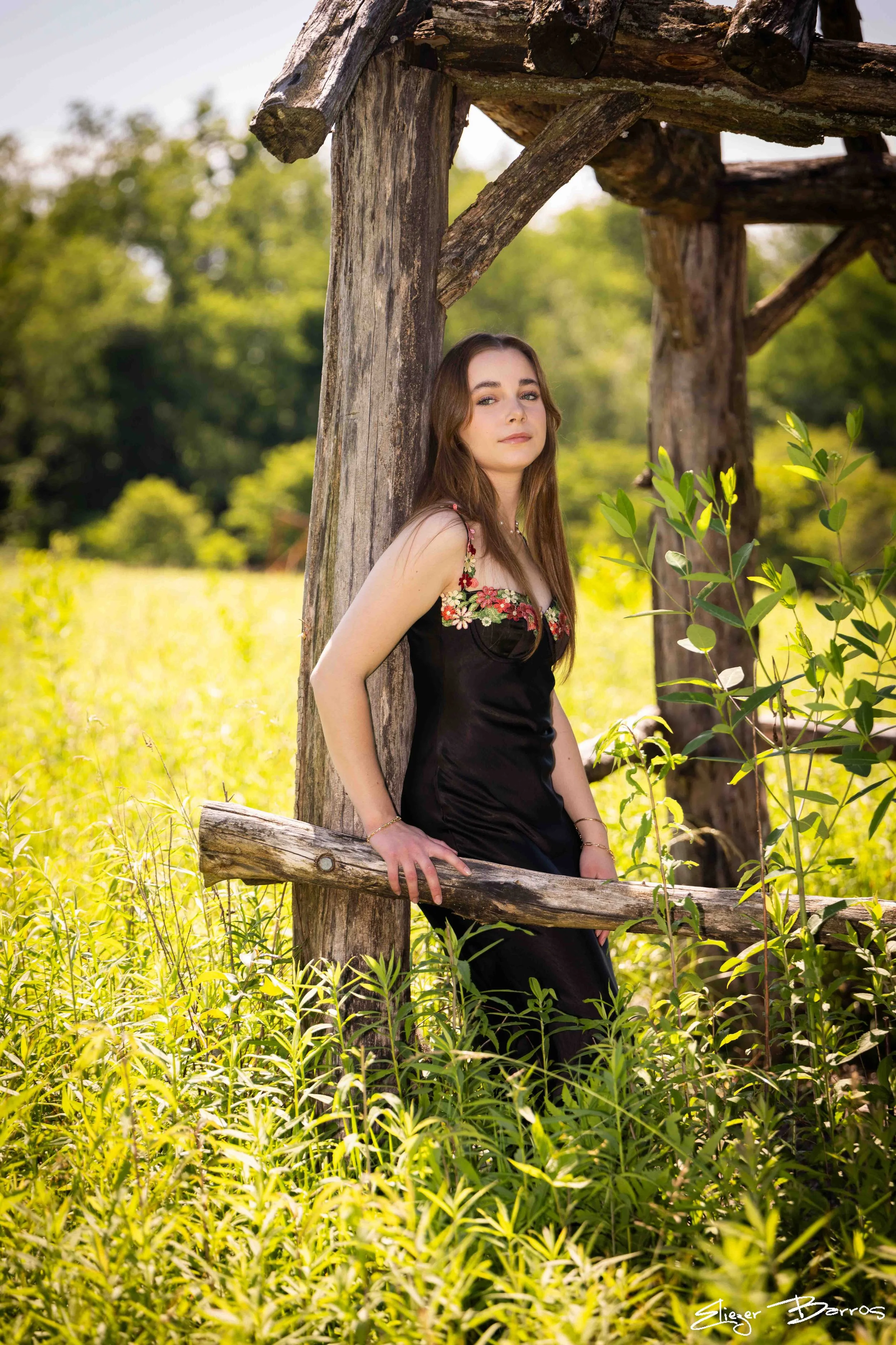 A young woman in a black dress with floral embroidery standing outdoors in a lush, green field by a rustic wooden fence. She leans against a large wooden post and gazes softly at the camera with a serene expression. The background is filled with vibr