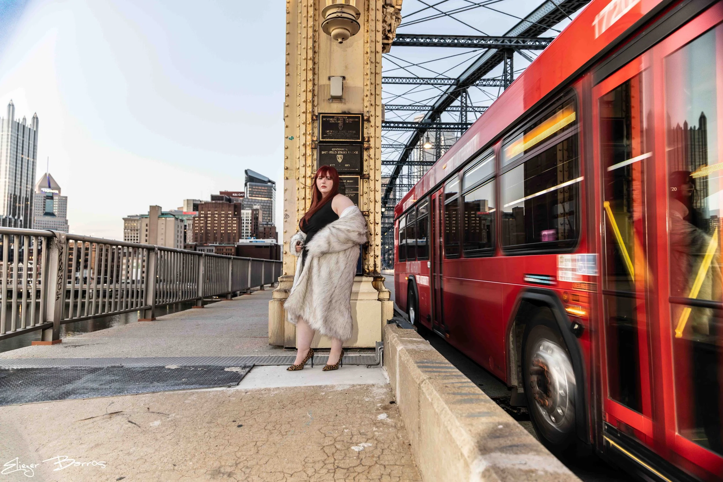 A woman with red hair standing on a city bridge near a red bus, wearing a black dress and a fur coat, with a city skyline and bridge structure in the background.
