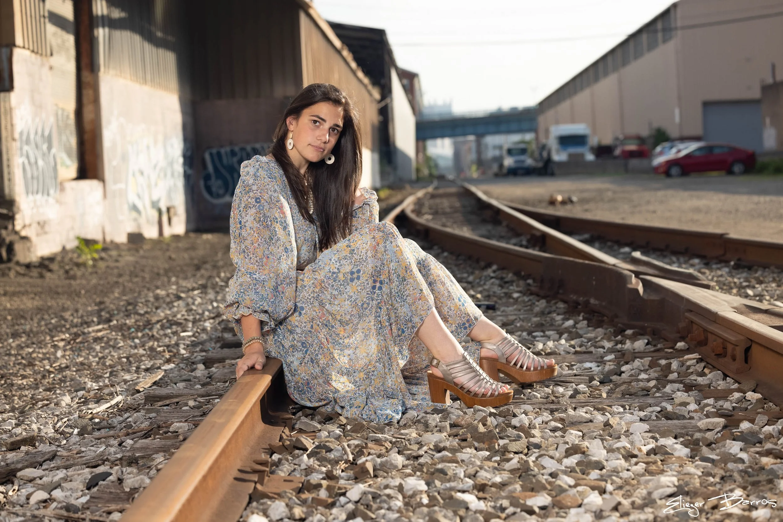 A young woman with long brown hair is sitting on a railroad track in an urban area, wearing a long floral dress and tan platform sandals, with a confident expression and large white earrings.