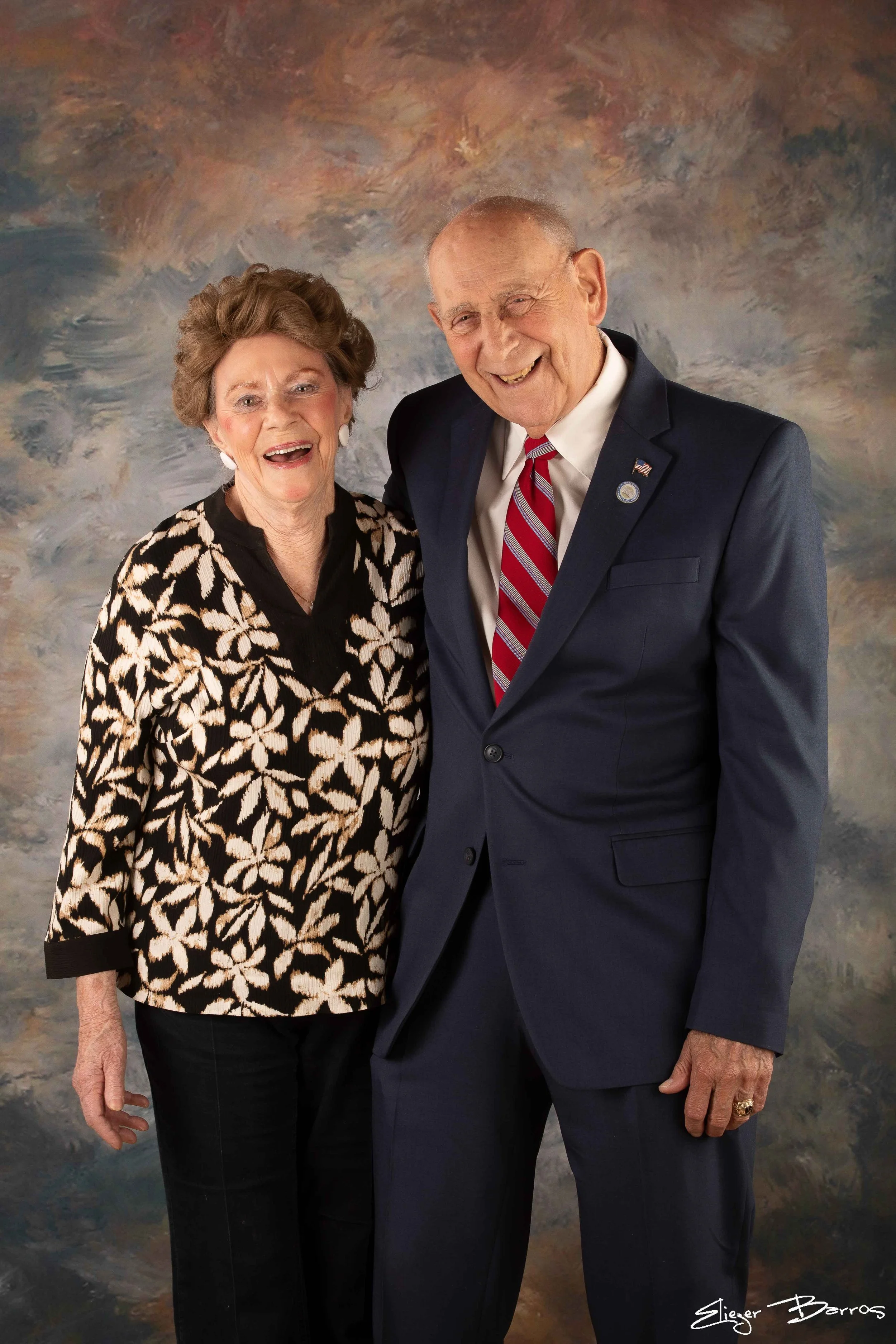 An elderly man and woman smiling and standing close together in front of a colorful, abstract background. The woman wears a black and white patterned top, and the man is dressed in a dark suit with a white shirt and red striped tie. The man has a pin