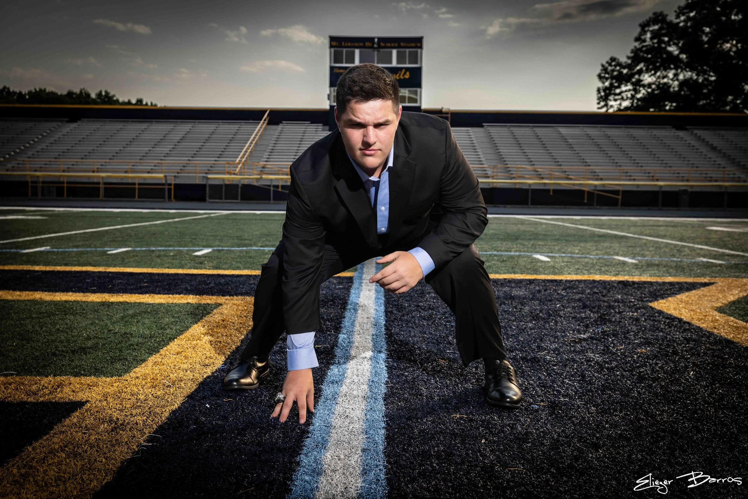 A young man dressed in a business suit crouching on a football field at the starting line, preparing to race, with empty bleachers and a scoreboard in the background at dusk.
