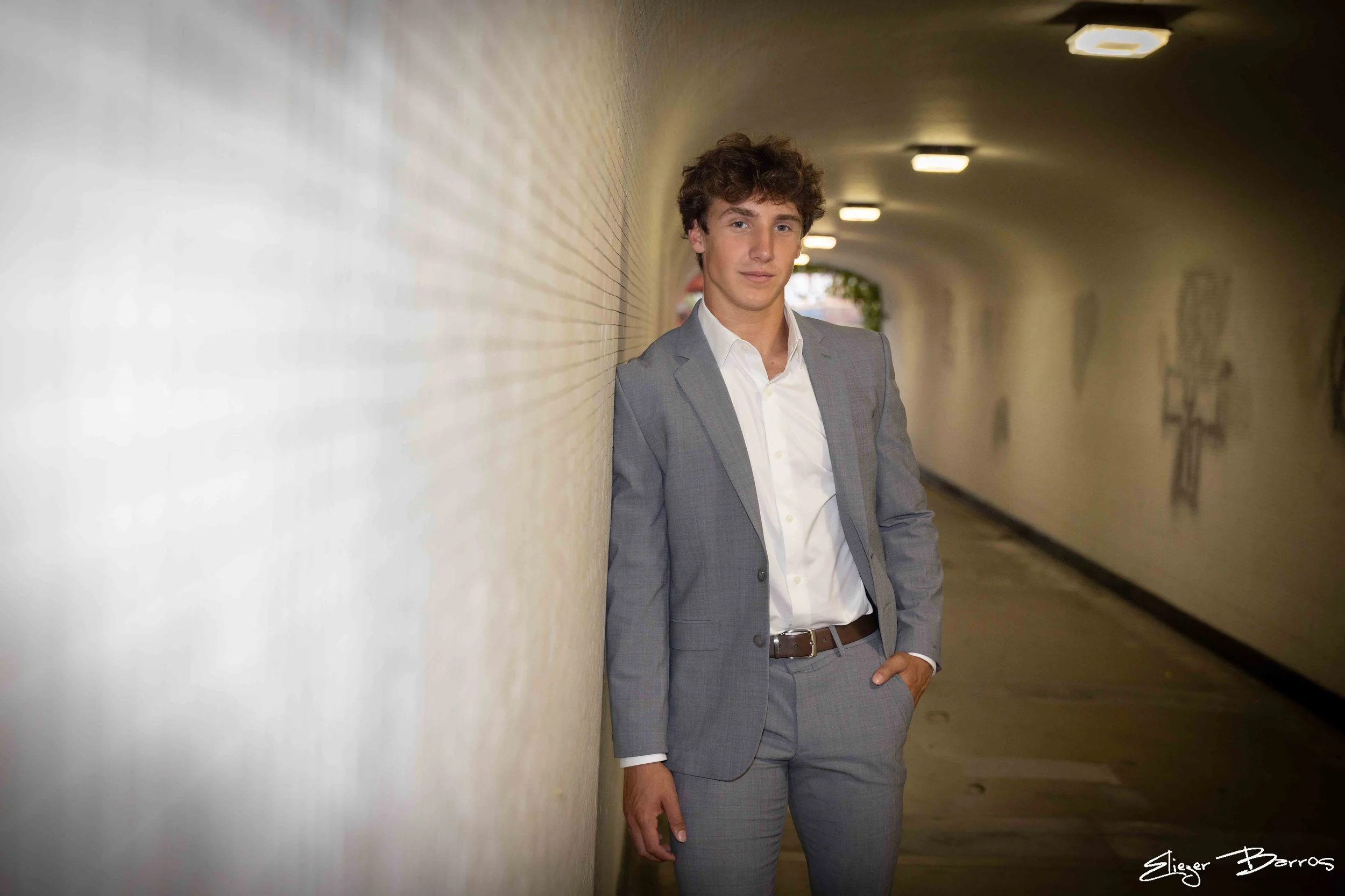 A young man in a light gray suit with a white shirt, standing in a tunnel or underpass with a brick wall on one side. He has curly brown hair and is looking towards the camera with his hand in his pocket.