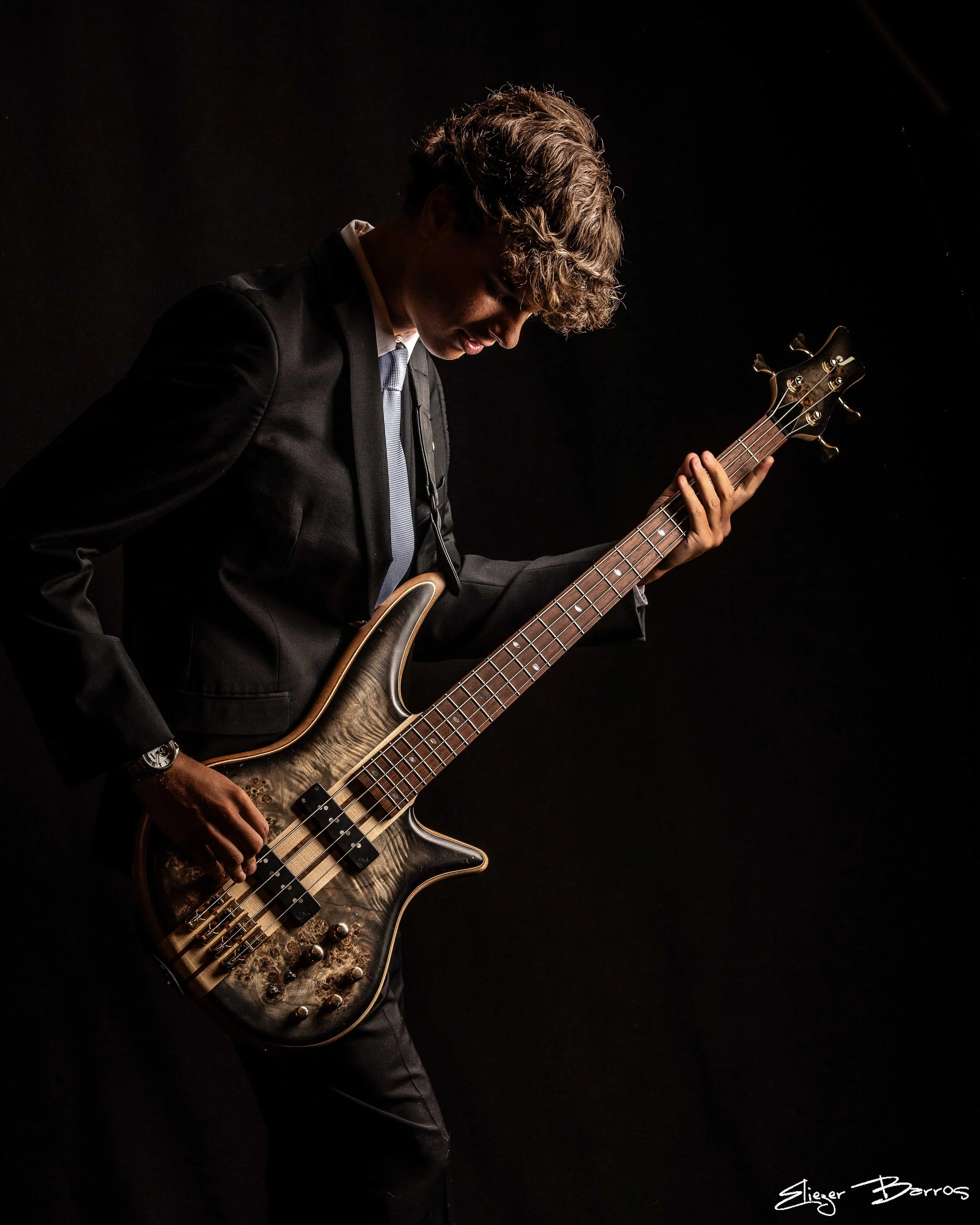 A young man in a black suit and blue tie is playing an electric bass guitar against a dark background. He has curly hair and is looking down at his instrument.