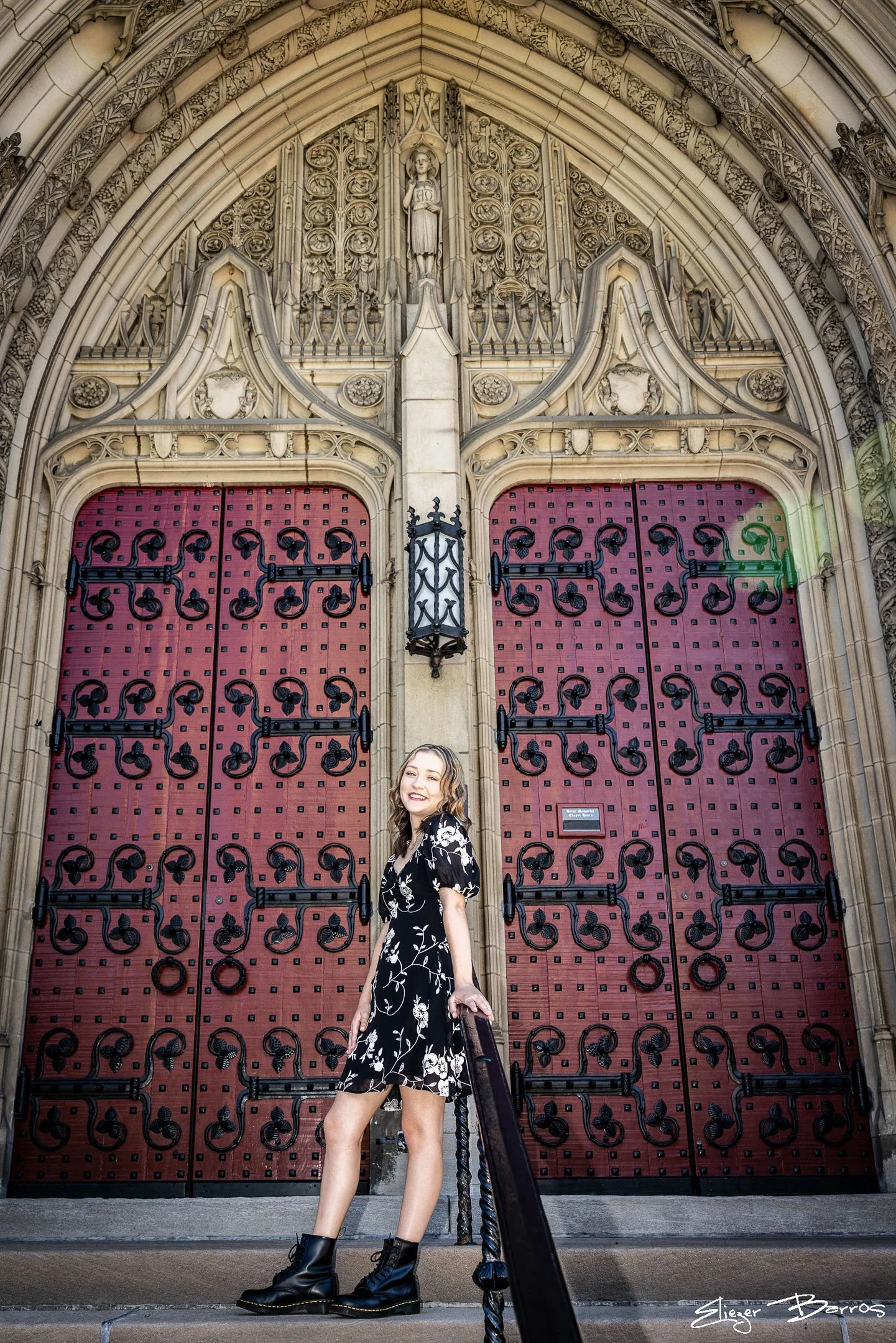 A young woman with wavy brown hair and a floral black and white dress standing in front of large red and black decorative metal doors and an ornate stone archway with detailed carvings and sculptures, including a statue of a woman above