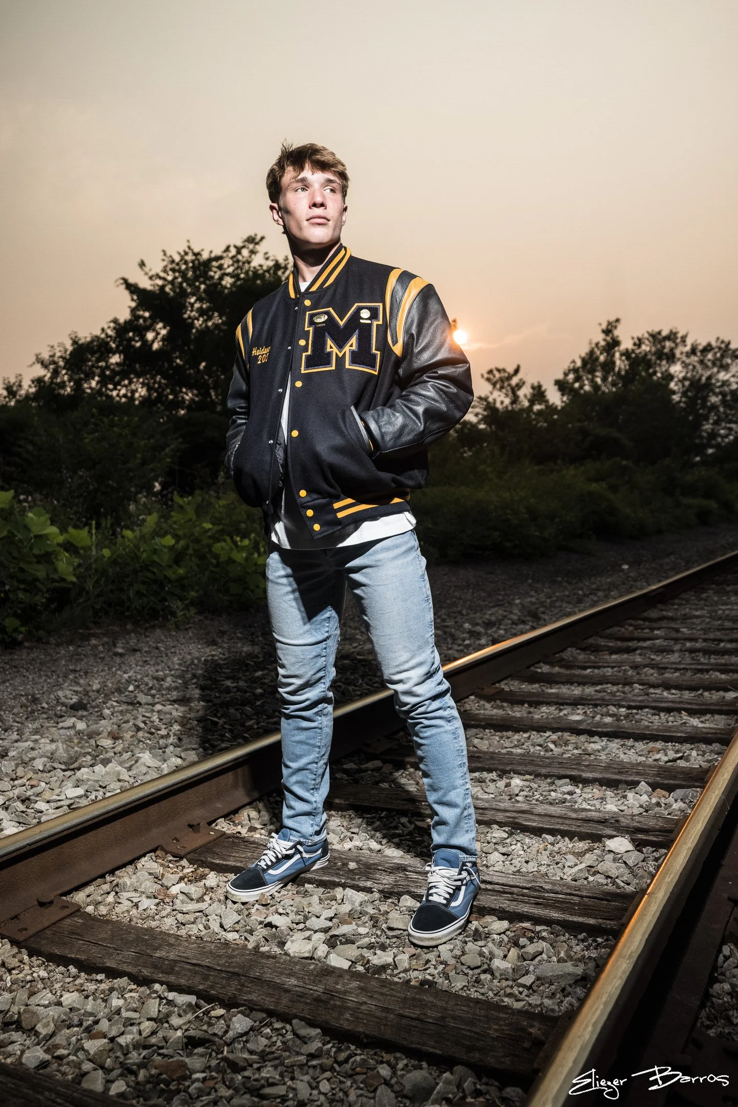 Young man in a blue and yellow varsity jacket, blue jeans, and sneakers standing on railroad tracks at sunset.