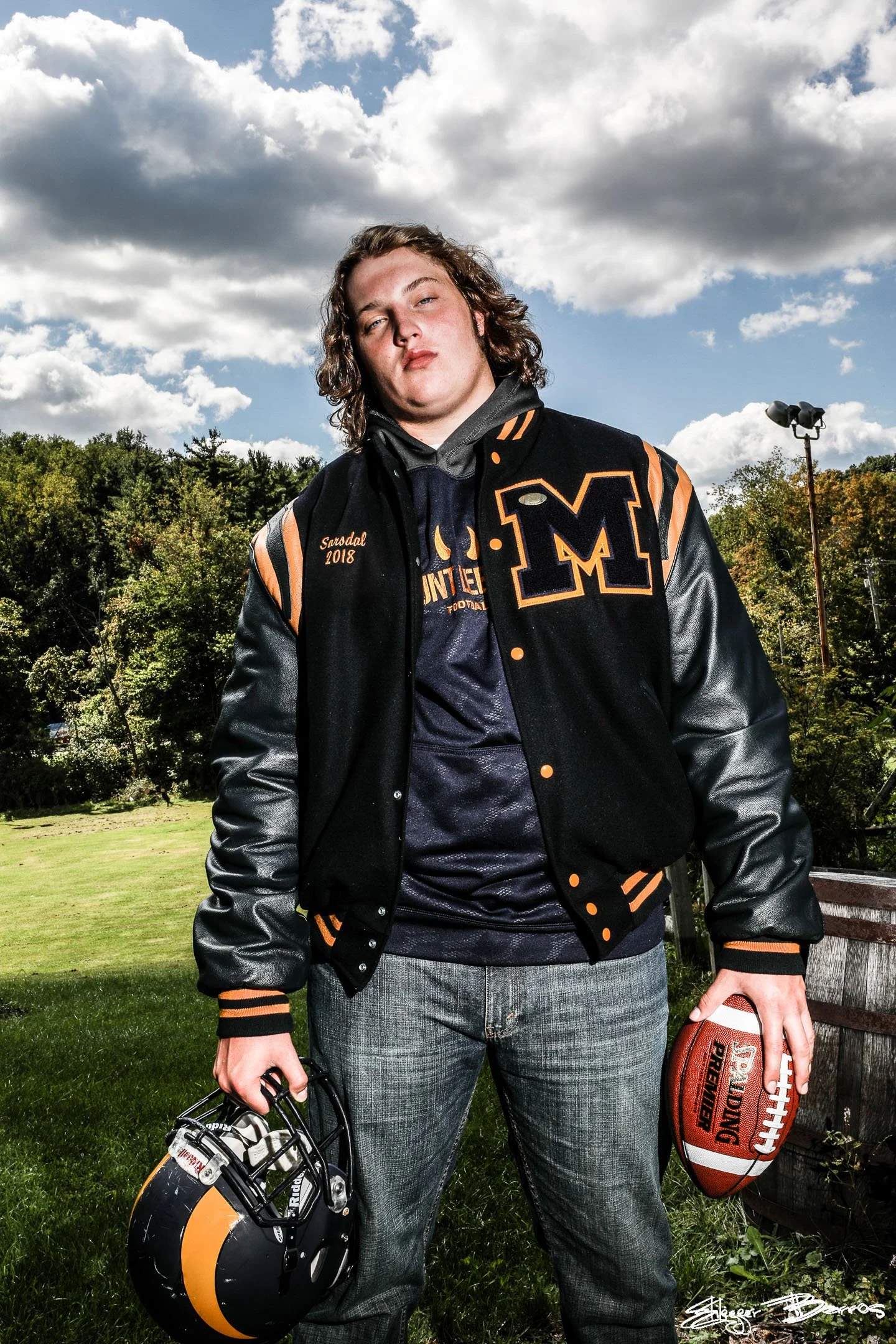 A young man standing outdoors on a grassy field, holding a football and a helmet, wearing a black and orange varsity jacket, with trees and a cloudy sky in the background.