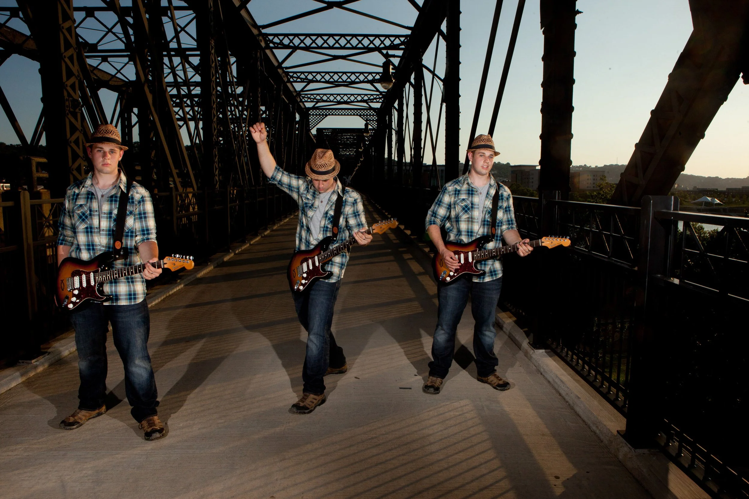 Three young men wearing plaid shirts, jeans, and straw hats are walking on a bridge at sunset, each holding an electric guitar.
