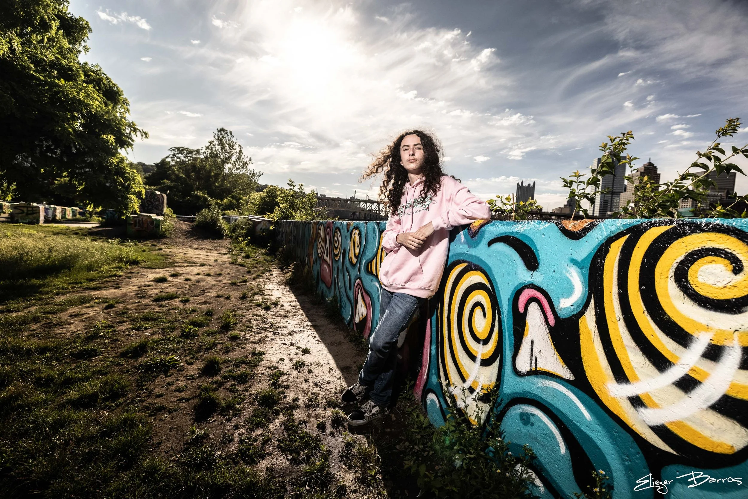 A young woman with curly hair standing outdoors next to a colorful graffiti-painted wall, with a city skyline and a large bridge in the background. The sun is shining through cloudy skies.