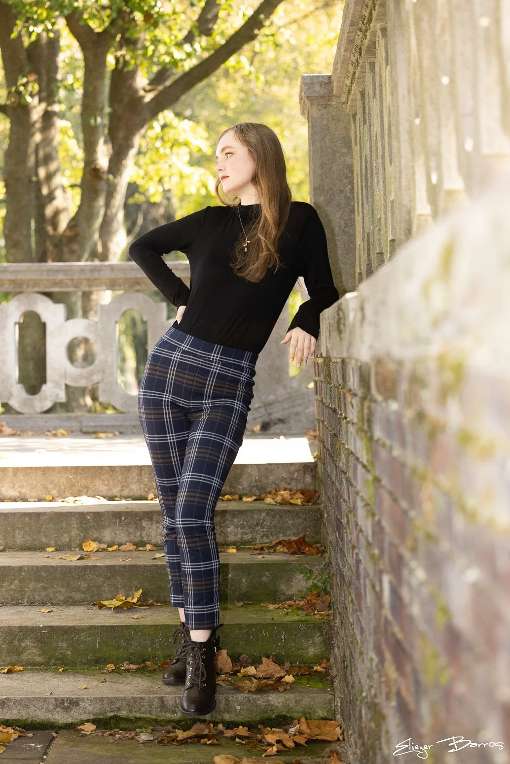 A young woman with long brown hair leaning against a stone wall outdoors in autumn, wearing a black long sleeve top, plaid pants, and black combat boots.