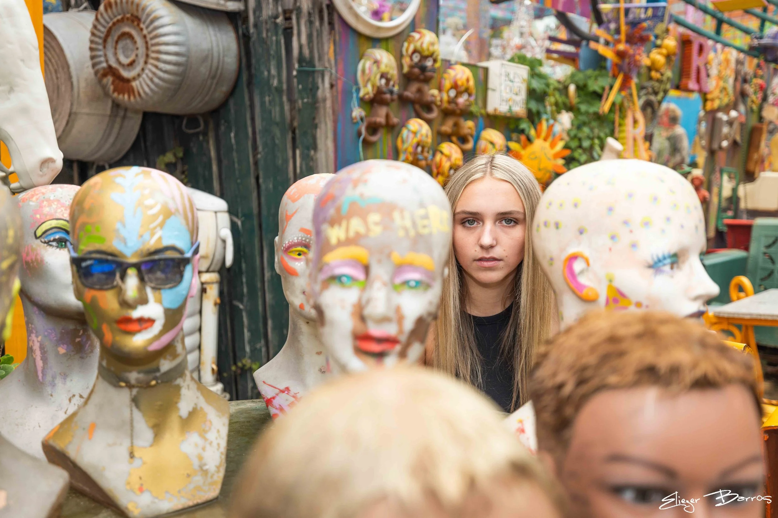 A young woman with blonde hair looking through a display of decorated mannequin heads at an outdoor market or shop. The mannequin heads are painted with various colorful designs. There are also various decorative items and artwork visible in the back