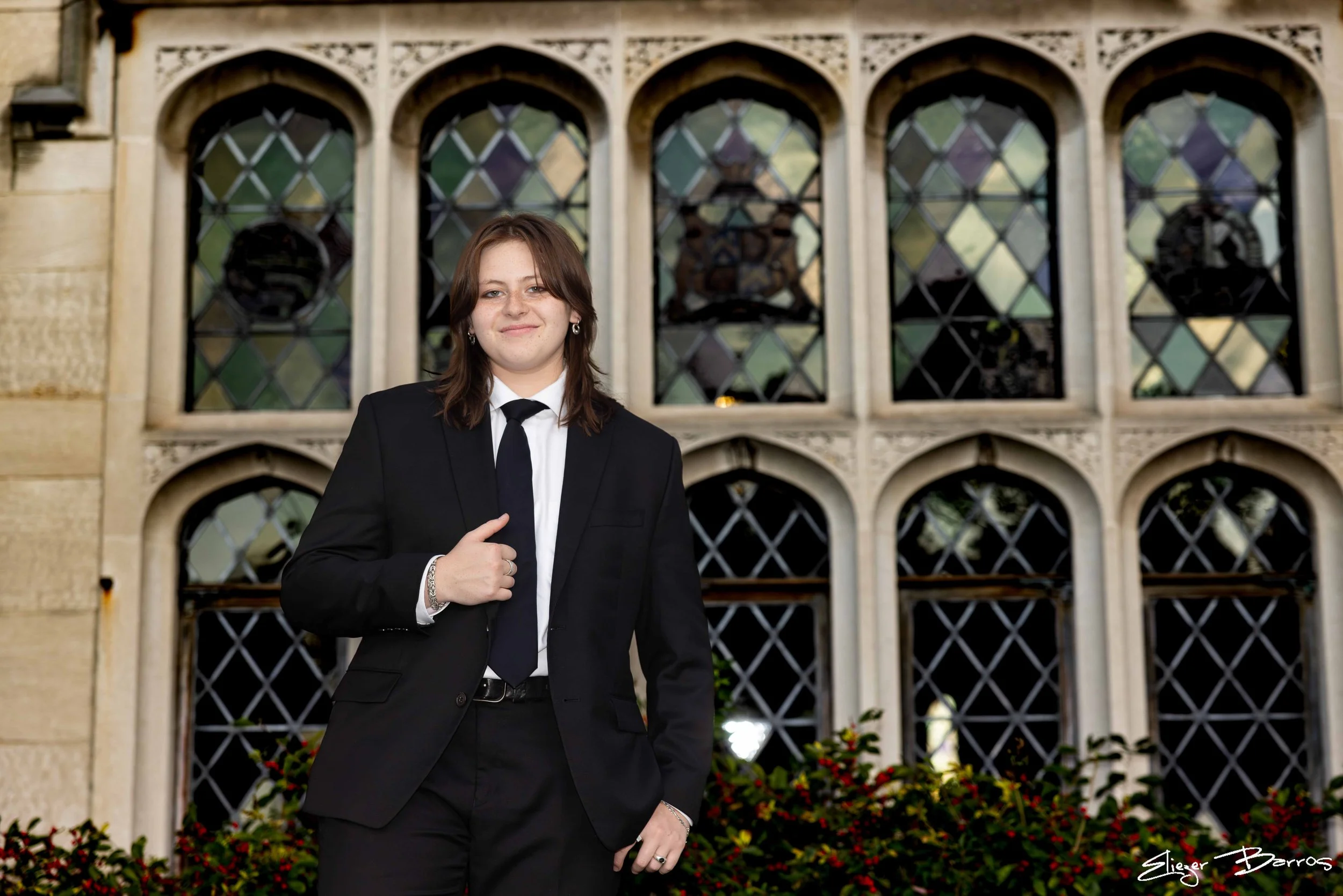 A woman in a black suit and tie standing in front of a stained-glass window of an old building, smiling with her right hand on her chest.