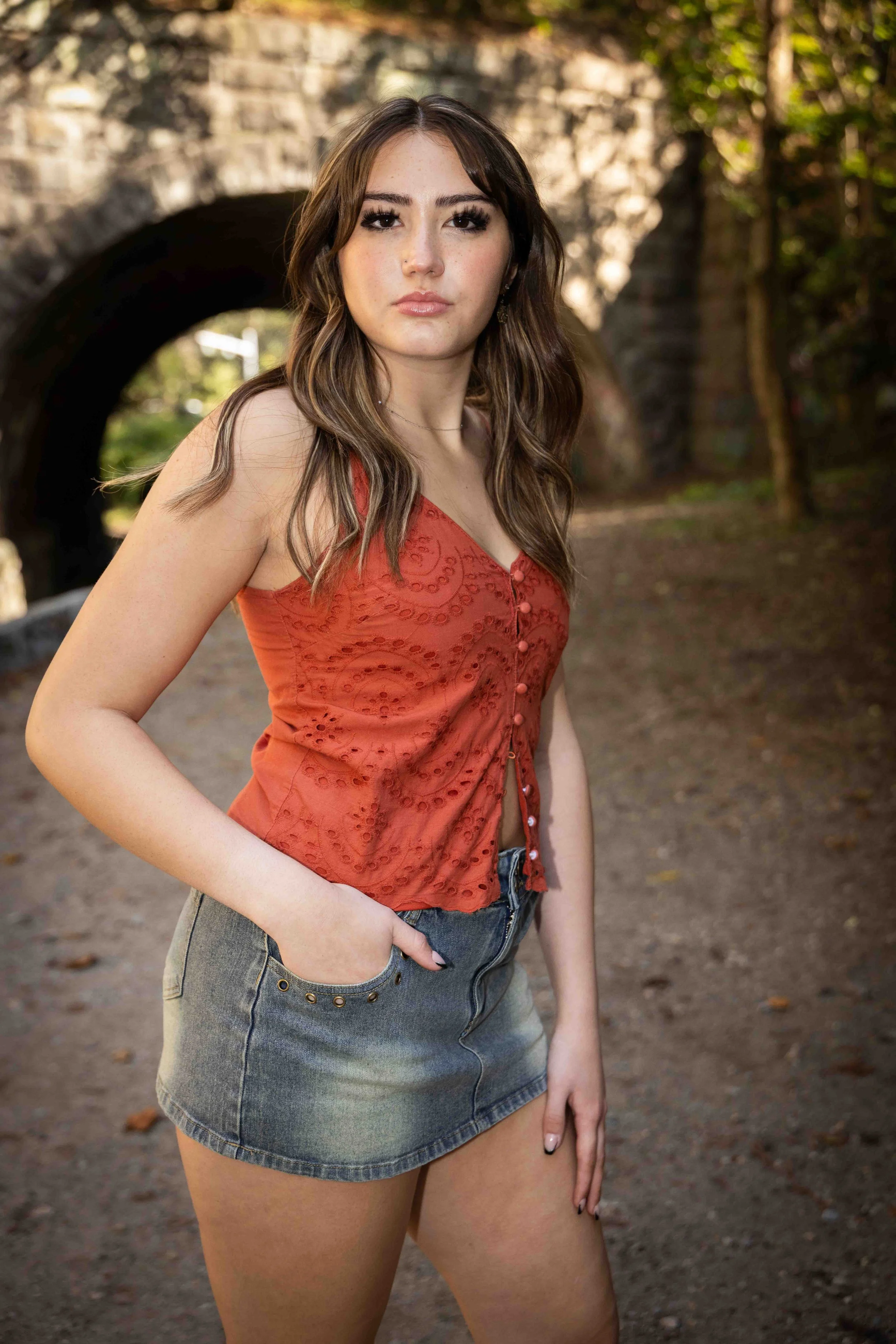 A young woman with long brown hair and wearing a red sleeveless top and denim shorts, standing outdoors near an arched stone bridge with trees and grass in the background.